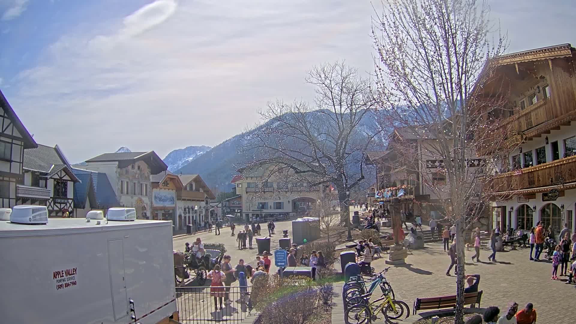A sunny day in a village square with European-style buildings, mountains in the background, and many people walking around.