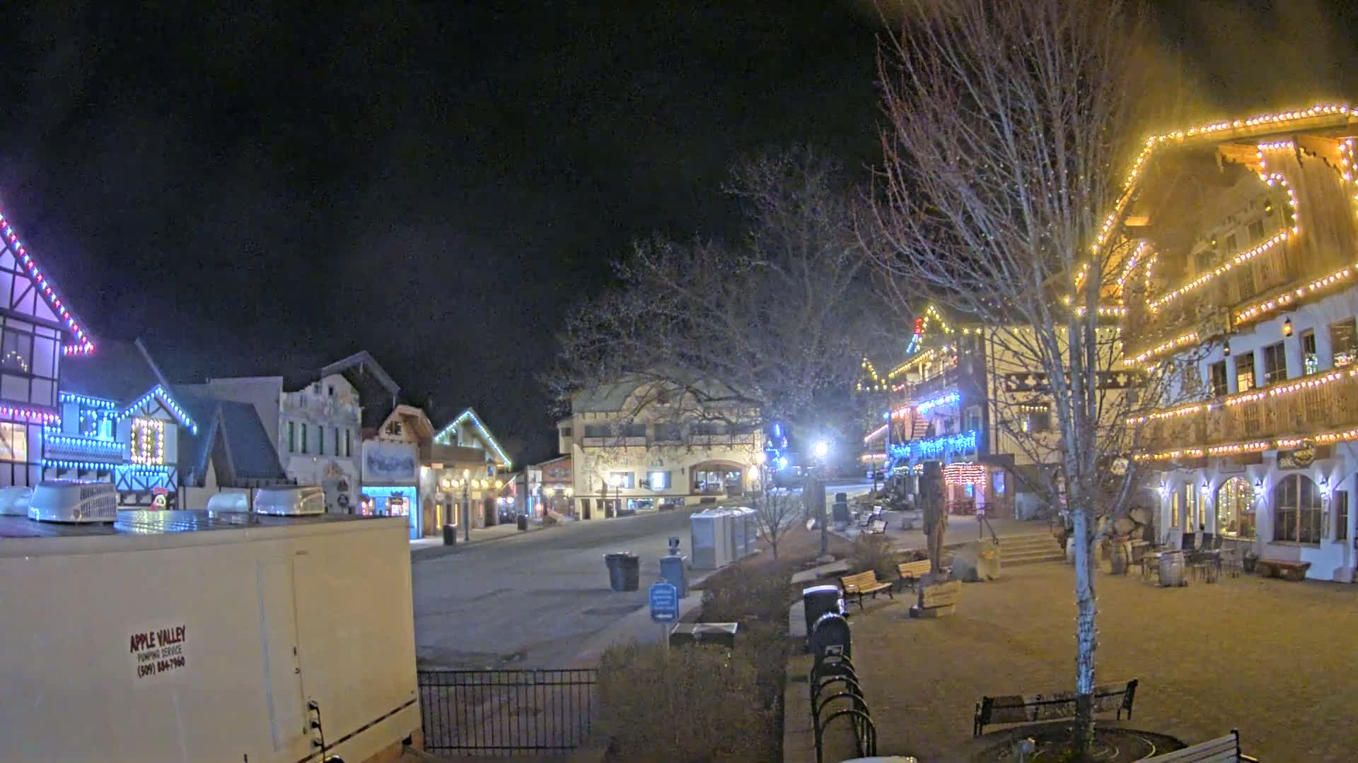 A nighttime scene of a town square with several buildings decorated with Christmas lights, bare trees, and empty benches.