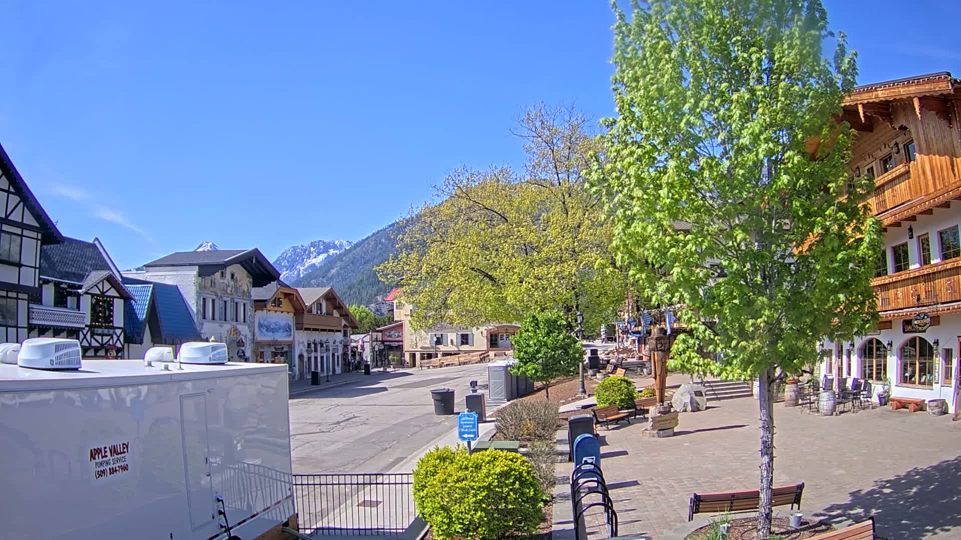 A mostly empty town square with European-style buildings and green trees under a clear blue sky, backed by snow-capped mountains.
