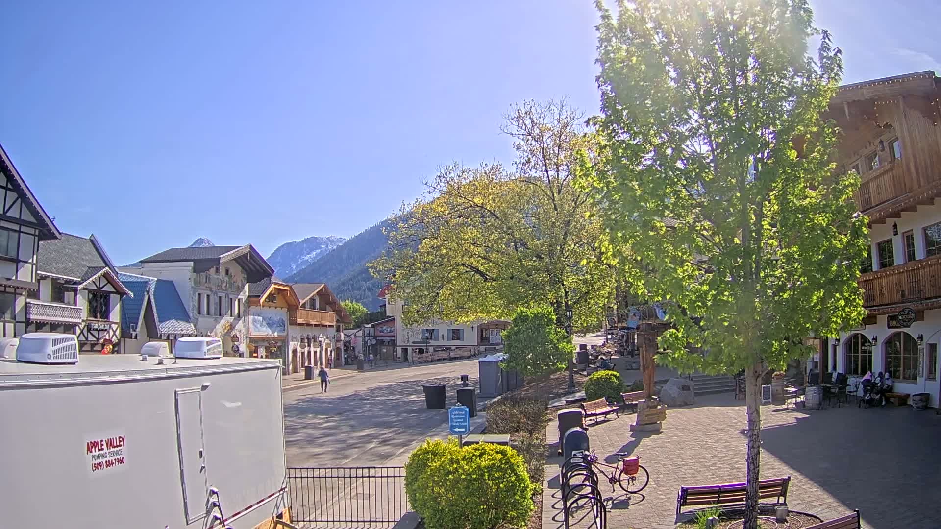 A sunny day reveals a village square with European-style buildings, lush greenery, and mountains in the background.