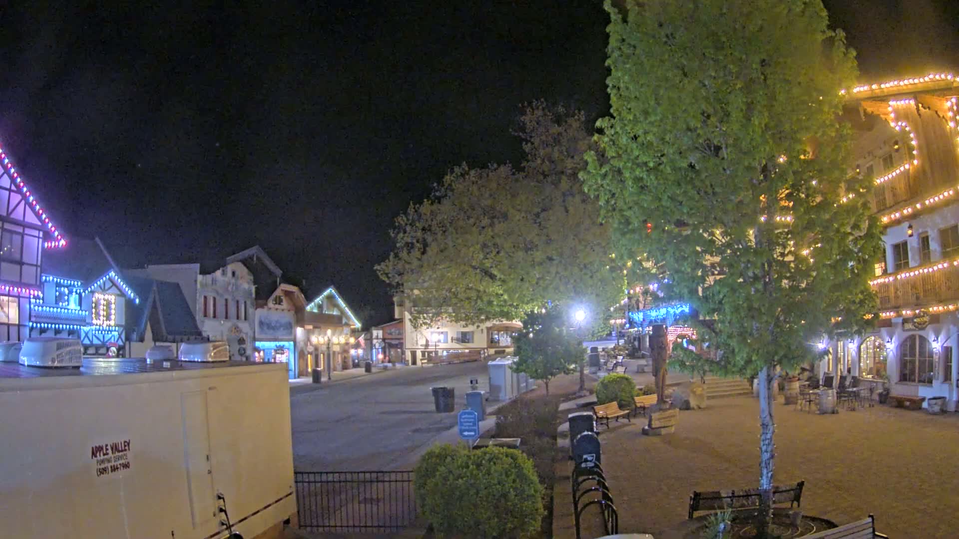 A nighttime scene of a town square with several buildings decorated with string lights,  trees, and benches.