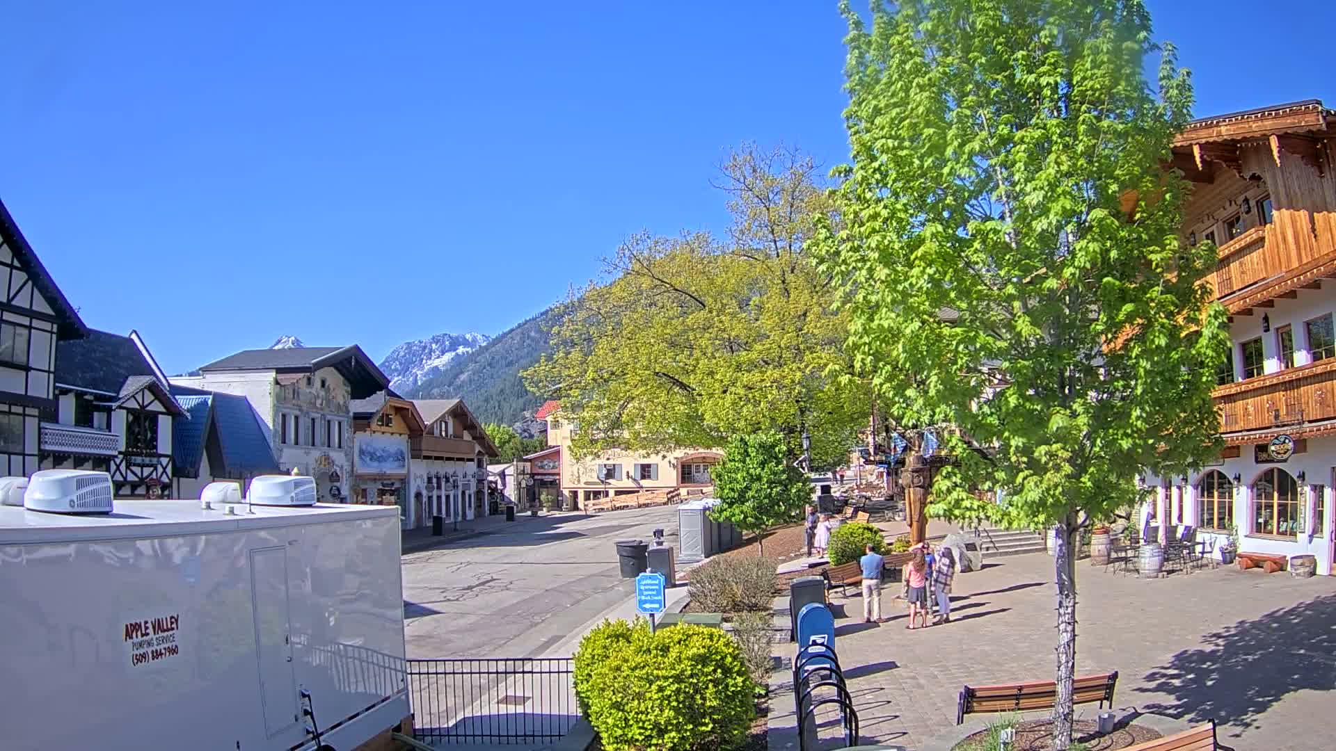 A mostly empty village street with several buildings of varying architectural styles, under a clear blue sky, with a mountain range in the background and green trees lining the street.