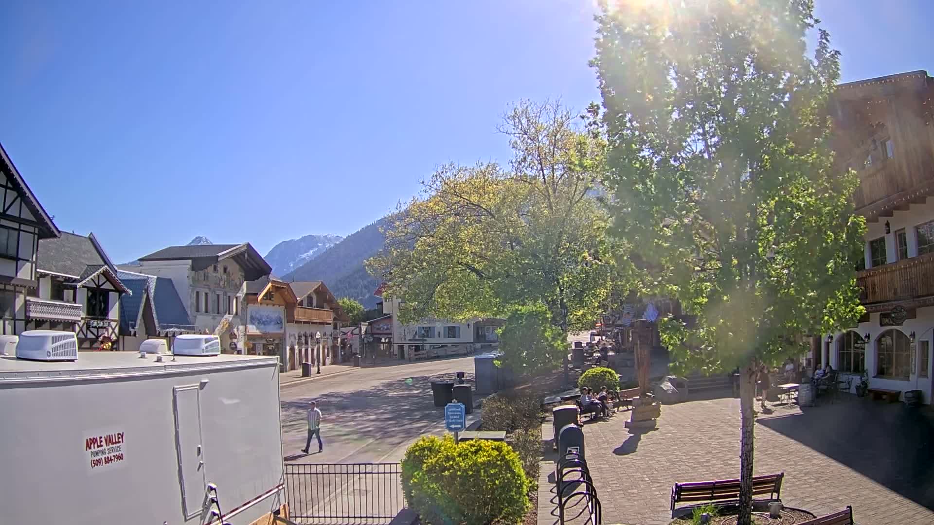 A sunny day in a quaint village, nestled at the foot of snow-capped mountains, shows a mostly empty street with people sitting at outdoor cafes and walking along a paved square.
