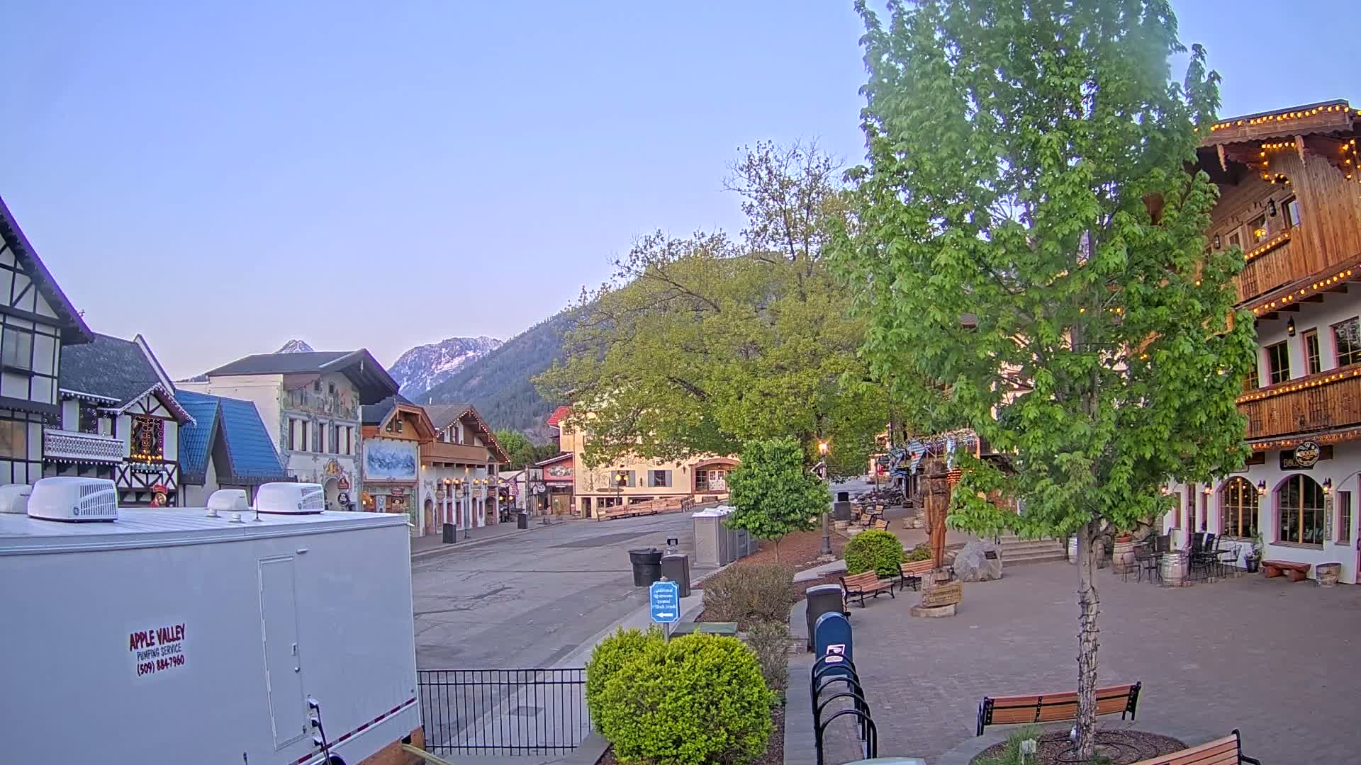 A mostly empty village square with European-style buildings, under a clear, twilight sky, is nestled at the base of snow-capped mountains.