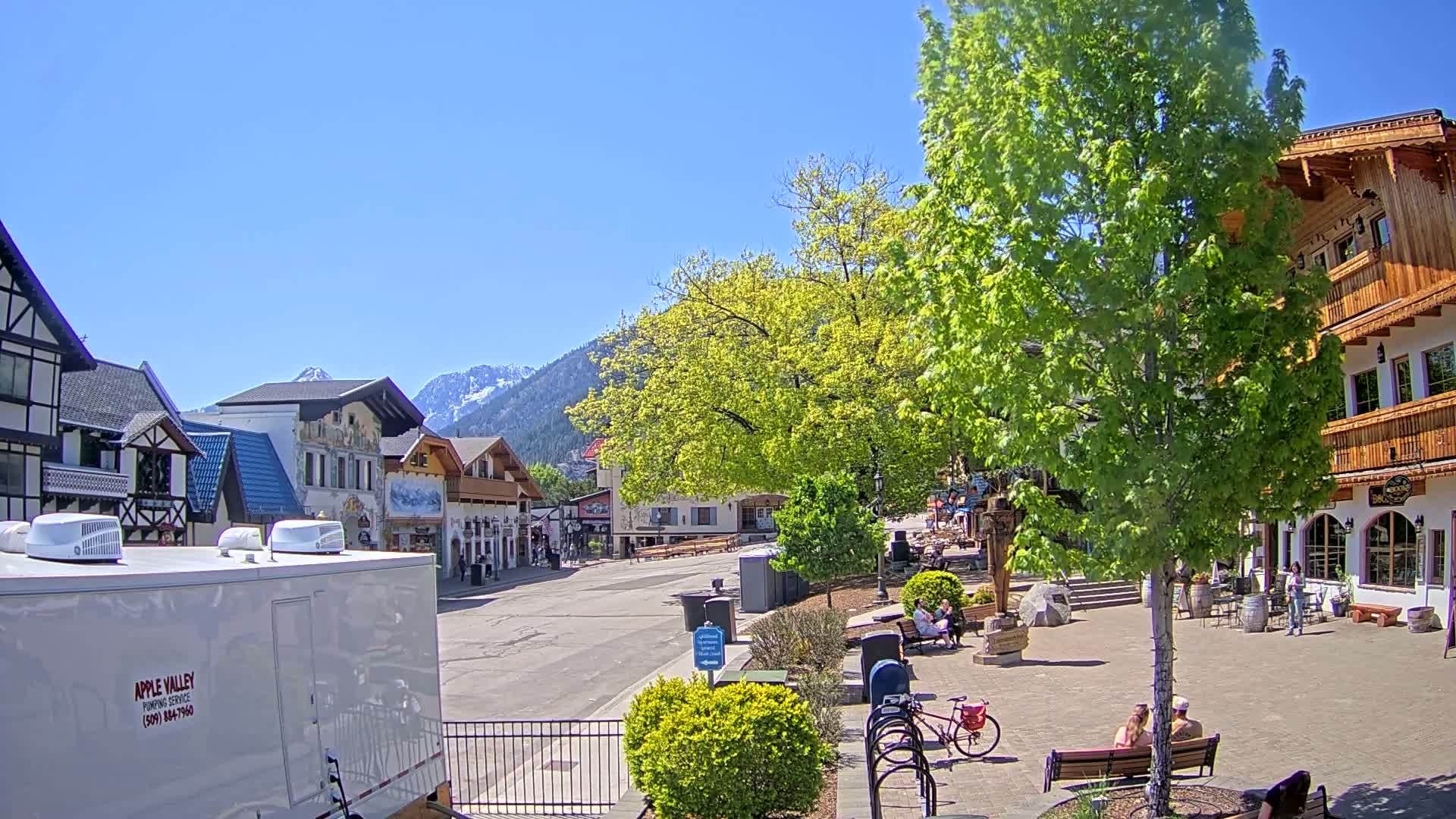 A village street scene with European-style buildings, green trees, and people relaxing outdoors on a sunny day in front of snow-capped mountains.