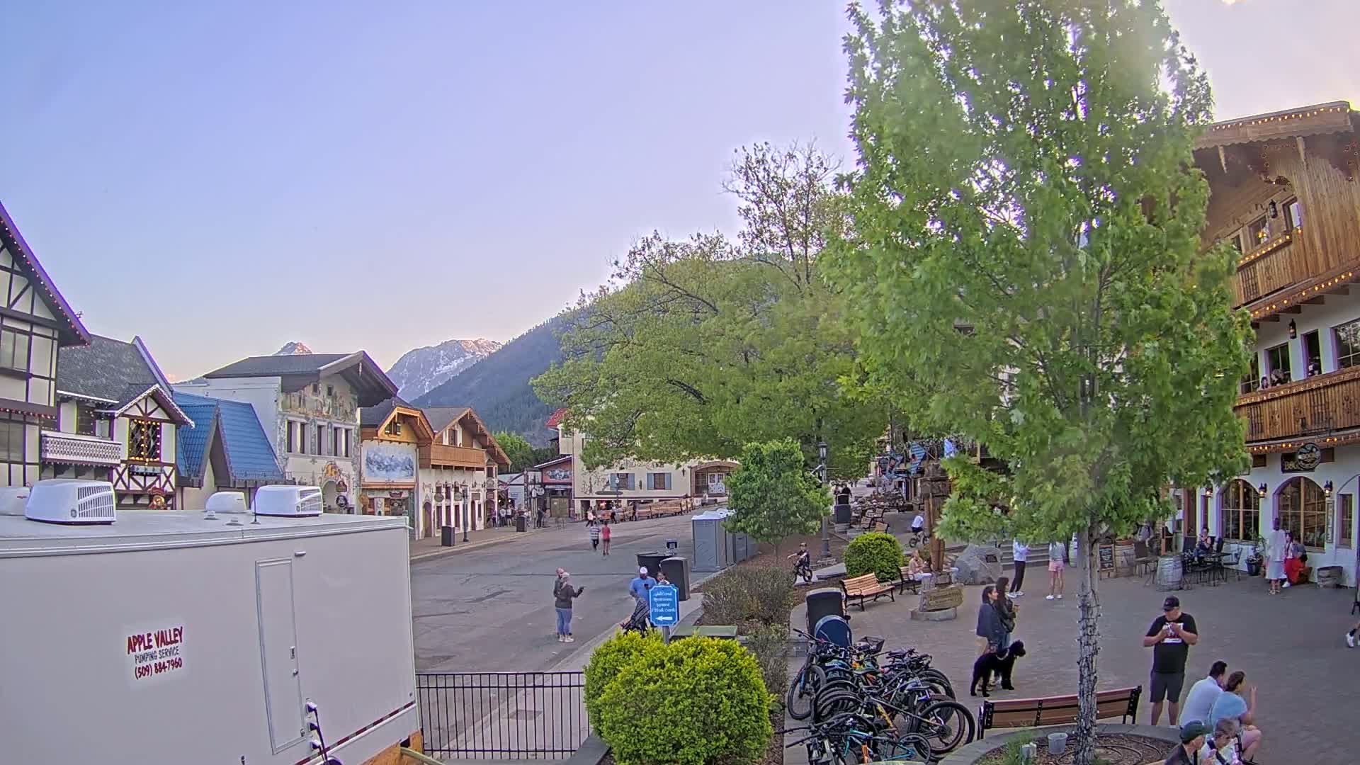 A mostly empty town square with several buildings in an alpine style, under a clear, light-blue sky, with mountains in the background and some people walking around.