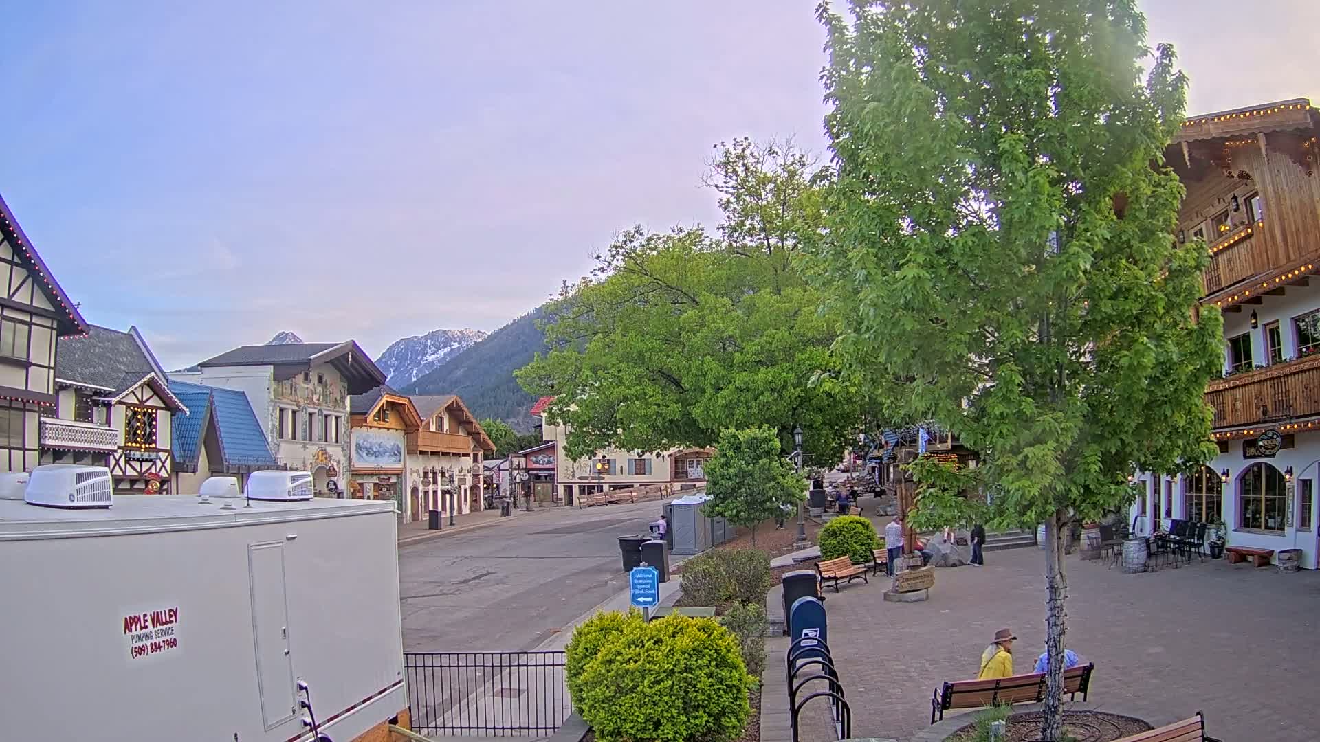 A mostly empty, European-style village street under a clear, light-blue sky, with mountains in the background and several people sitting on benches.