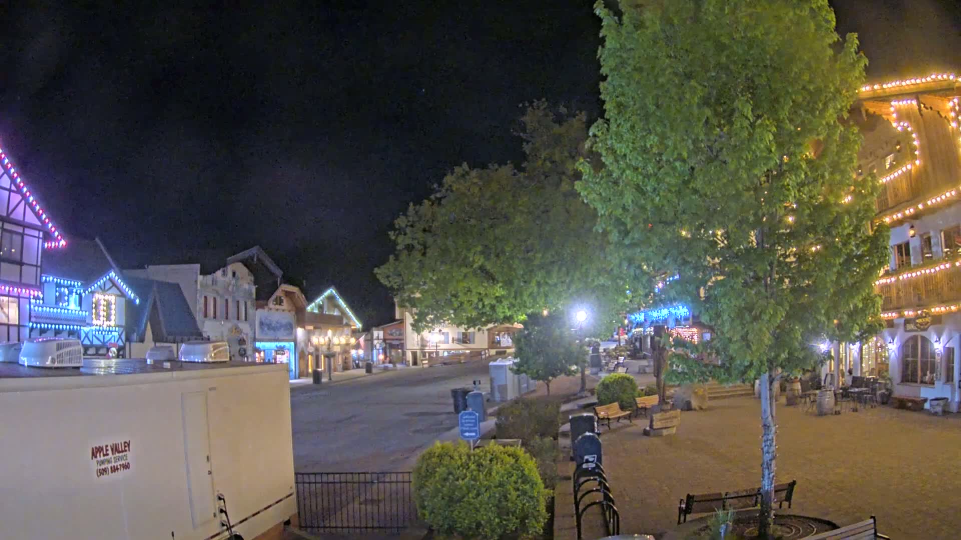 A nighttime street scene shows a row of festively lit buildings, with a large tree in the foreground and benches scattered throughout a paved plaza.