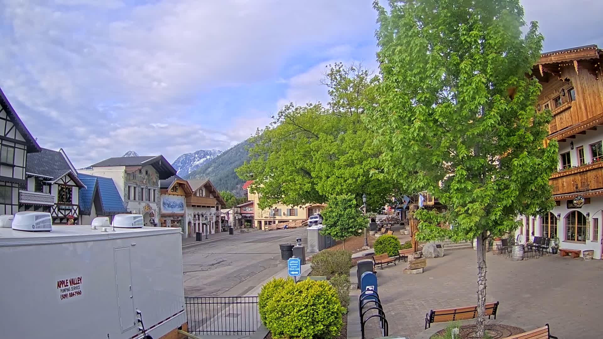 A partly cloudy day reveals a village square with European-style buildings, lush green trees, and snow-capped mountains in the distance.