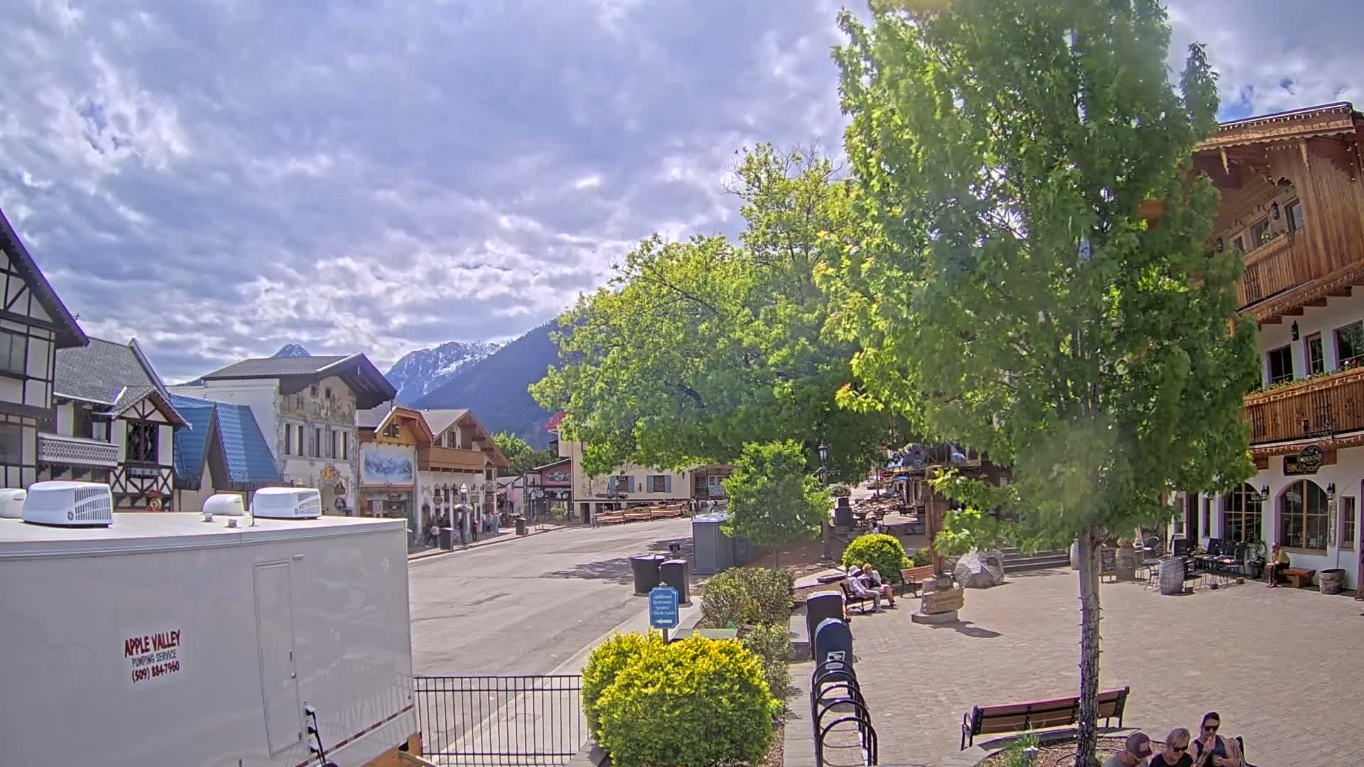 A partly cloudy day reveals a village square with several people seated near buildings of European architectural style, backed by snow-capped mountains.