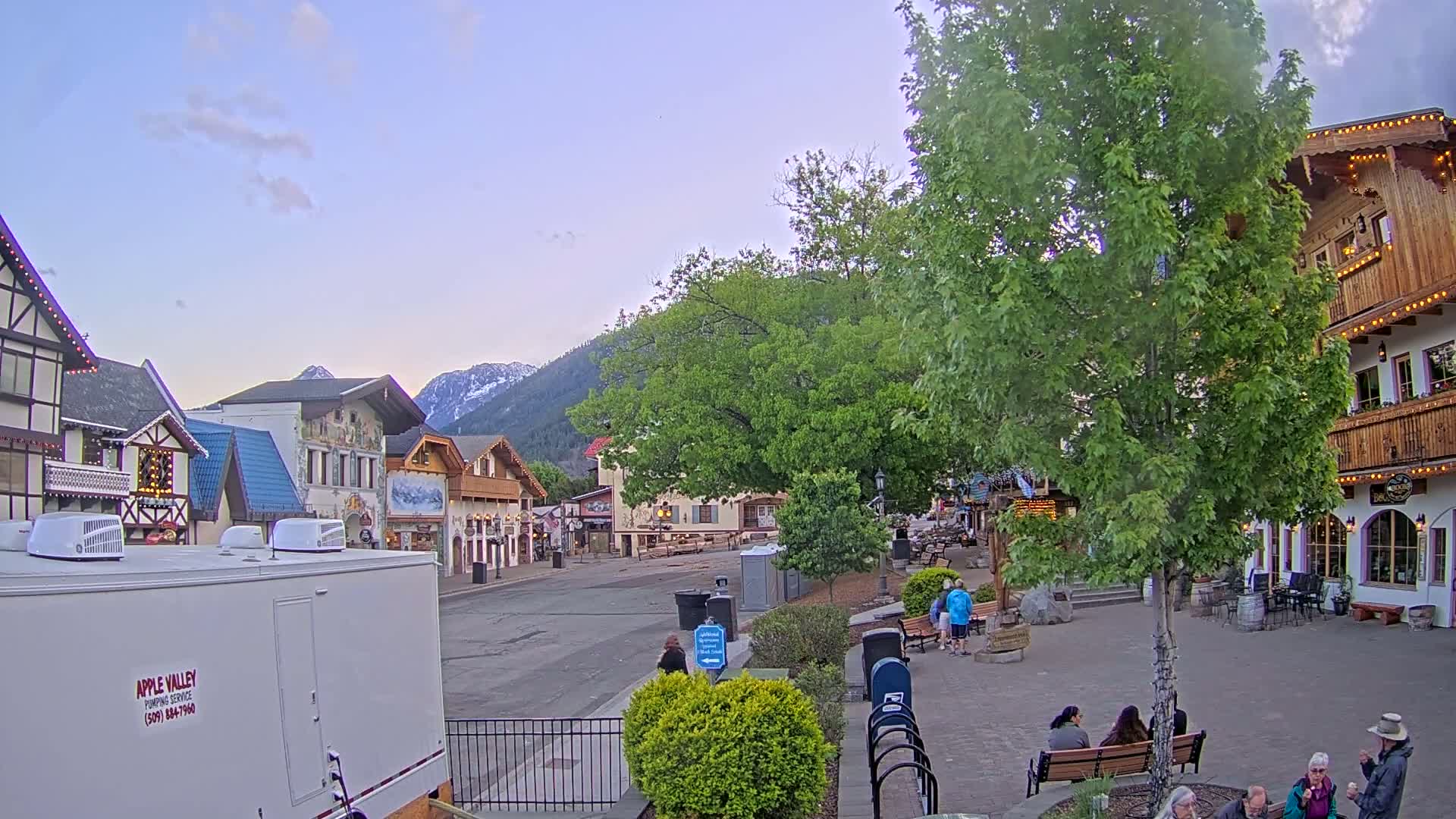 A quaint, European-style village street under a mostly clear, twilight sky is shown, with several people sitting on benches and walking, and mountains in the background.