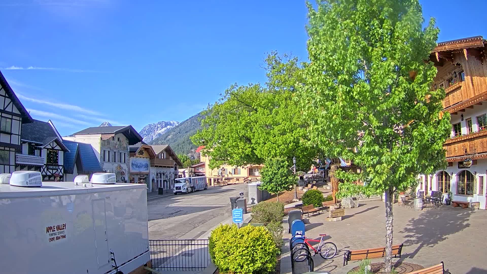 A village street scene on a sunny day, with snow-capped mountains in the background, features buildings in European architectural styles and lush green trees.