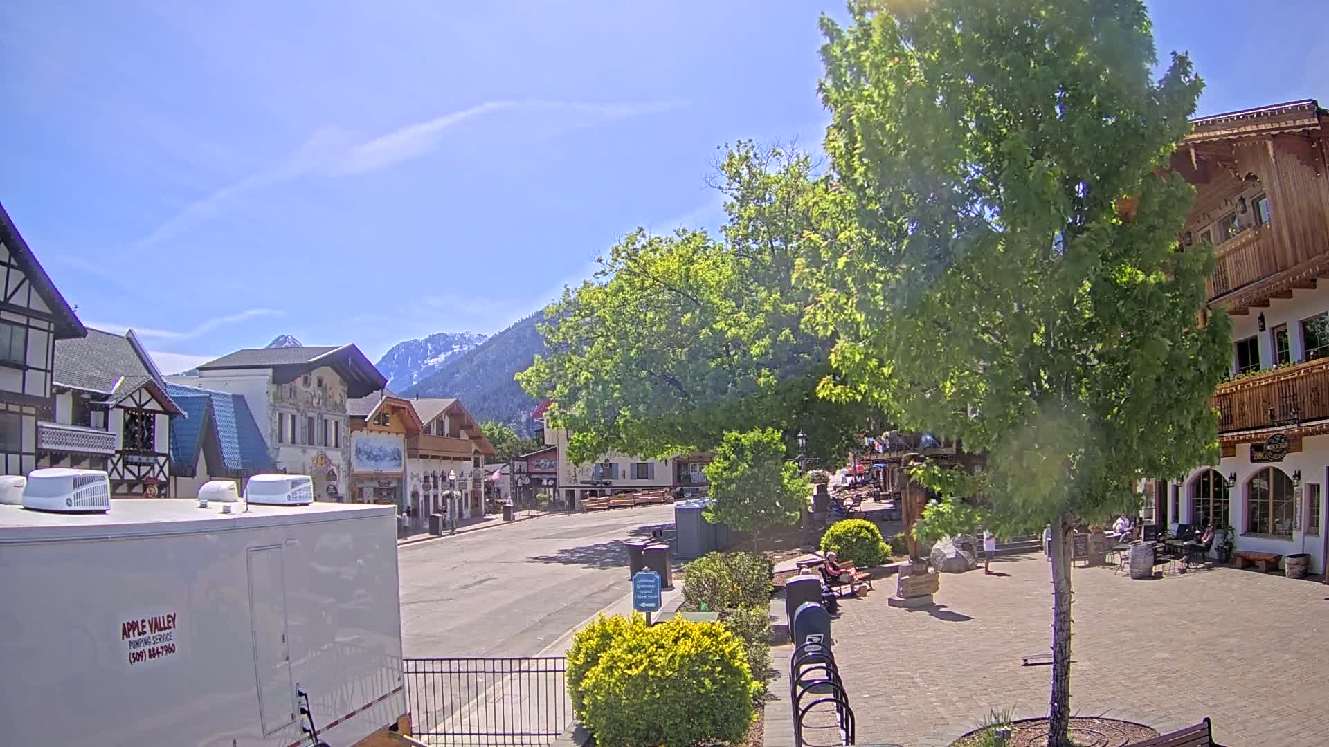 A sunny day reveals a town square with several buildings in a European architectural style, nestled at the base of snow-capped mountains.
