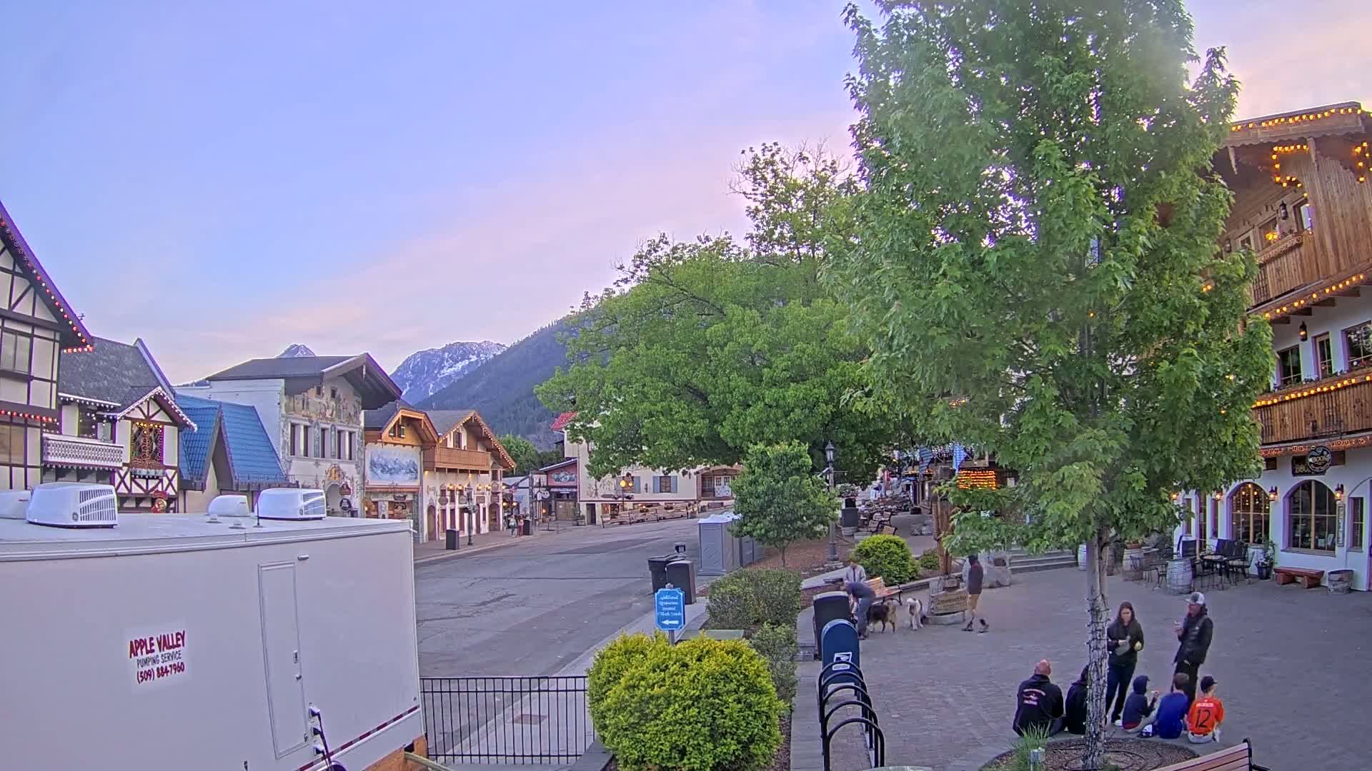 A quaint village street scene under a pale, dusk sky features several European-style buildings, lush green trees, and a few people gathered in a plaza.