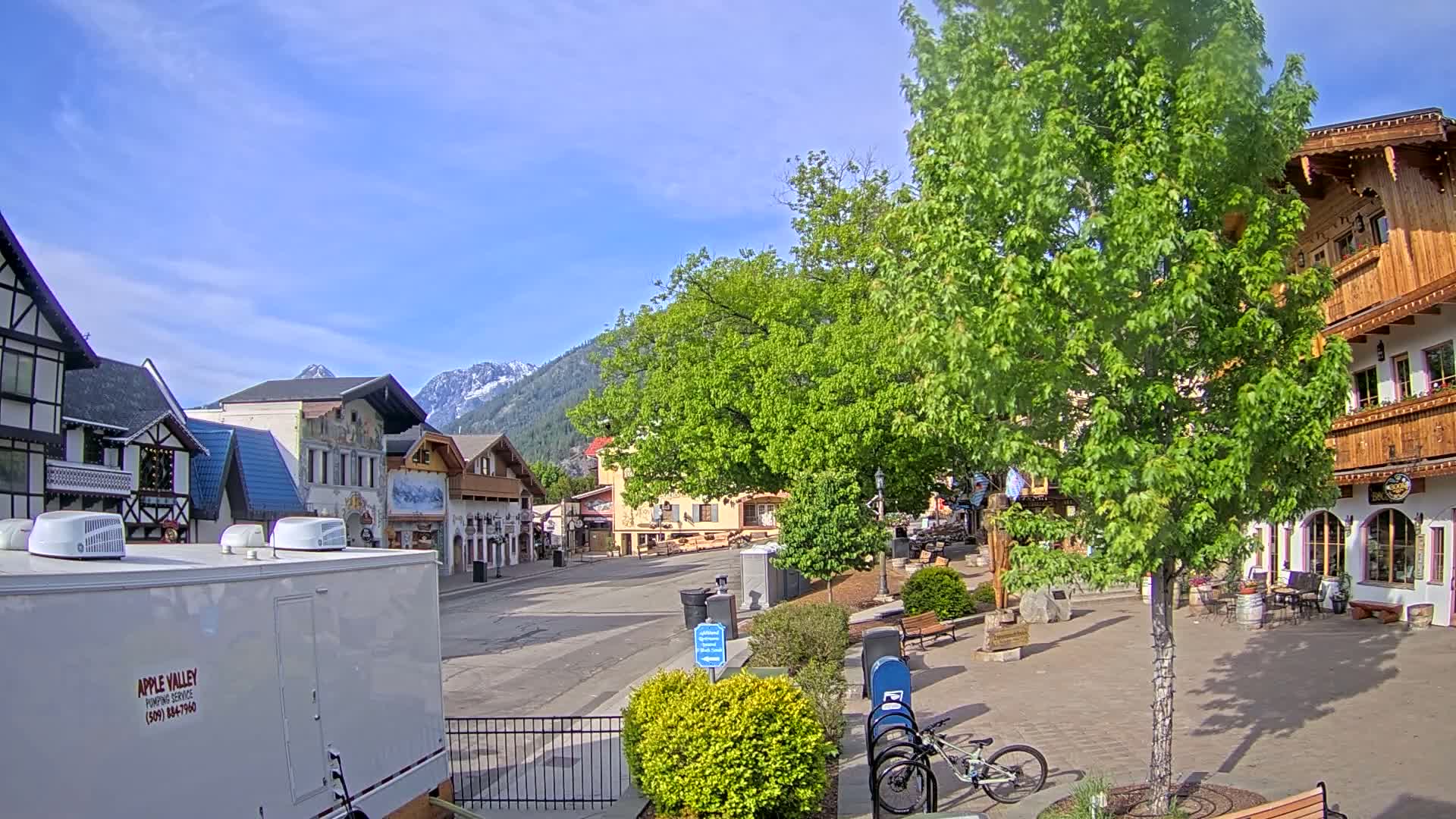 A sunny day reveals a quaint village street lined with European-style buildings, nestled against a backdrop of snow-capped mountains.