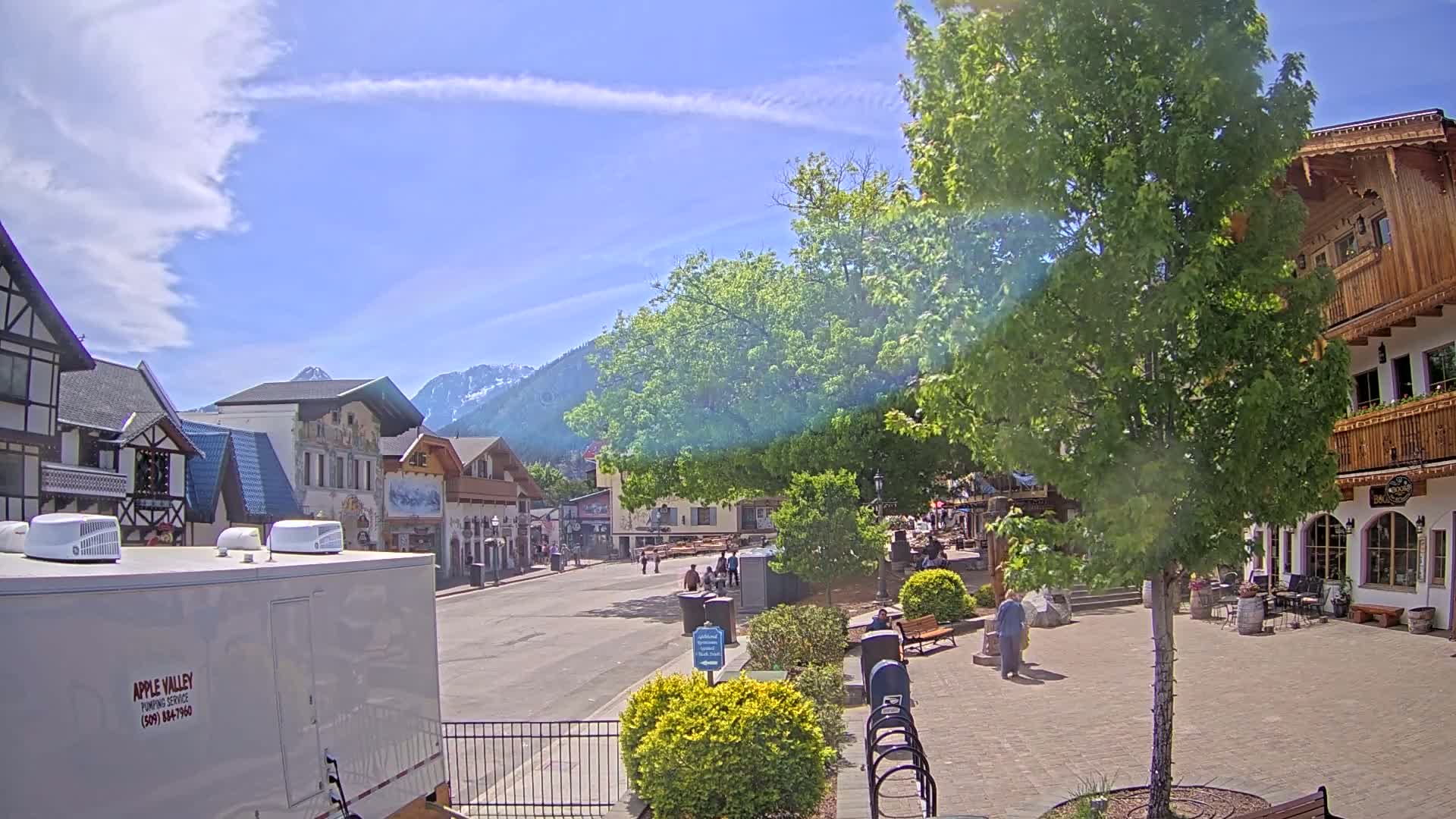 A partly sunny day reveals a village square with European-style buildings, a large tree, and mountains in the background.
