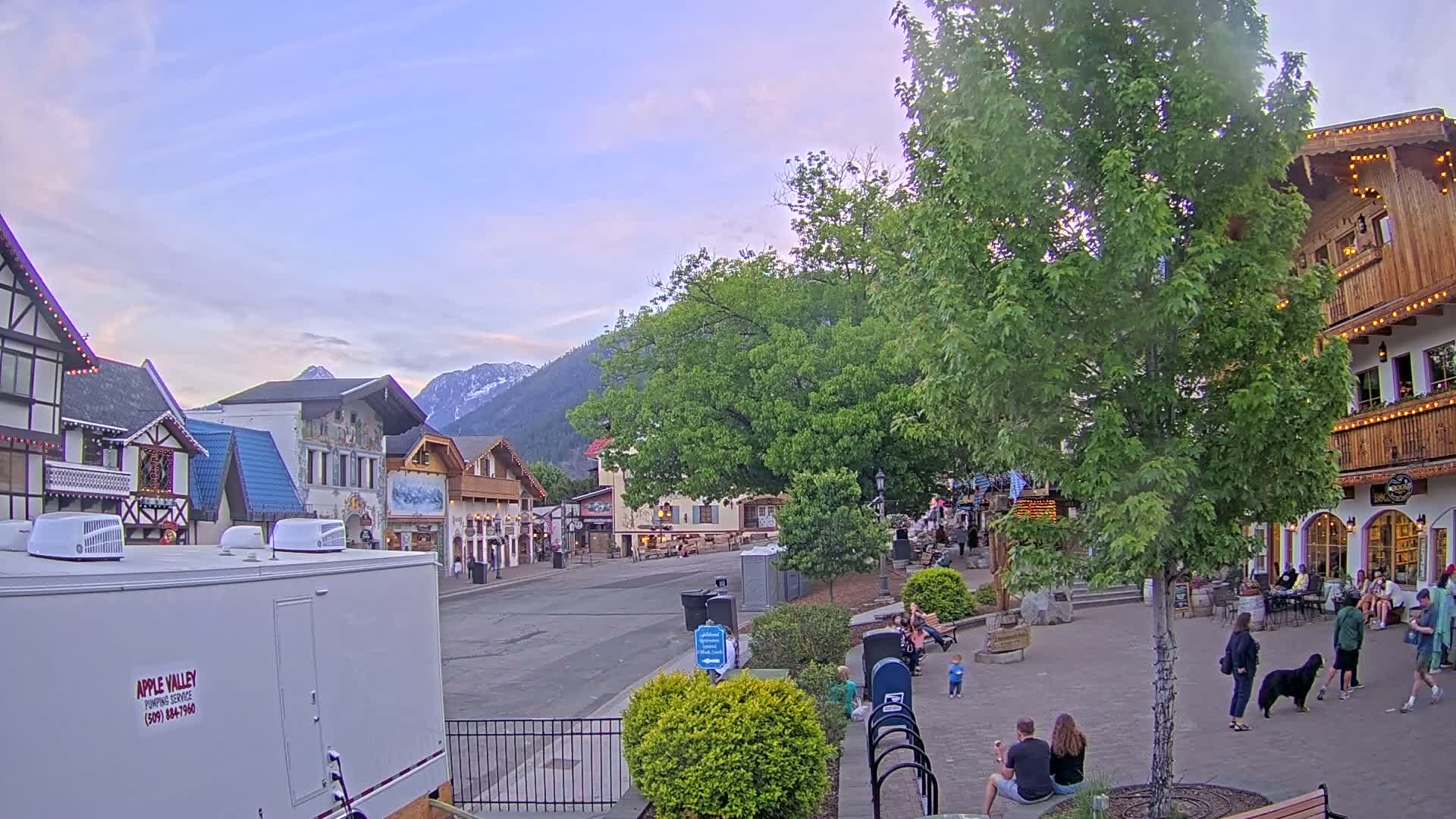 A quaint town square with European-style buildings, under a partly cloudy twilight sky, is surrounded by lush green trees and mountains in the background.