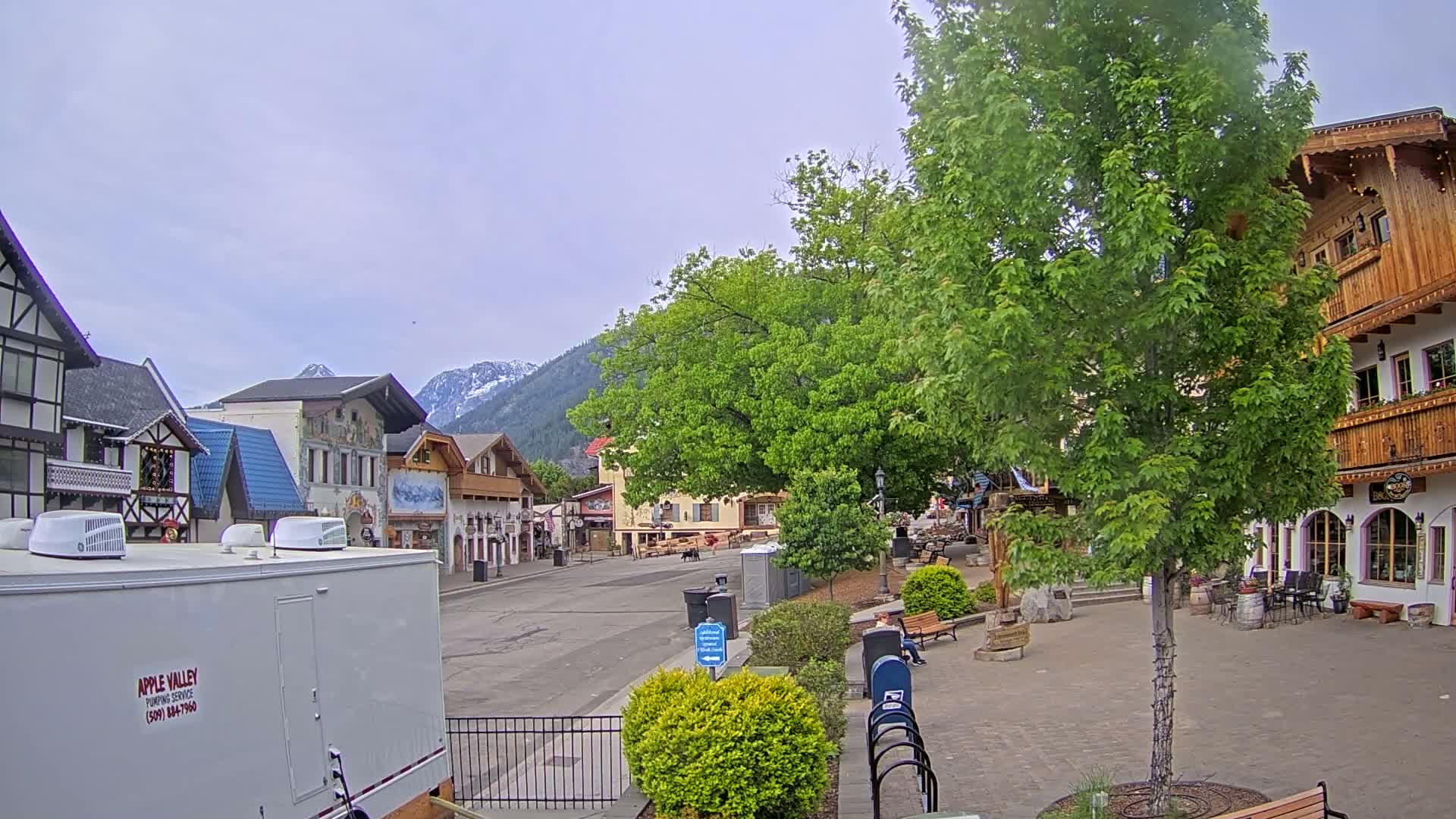 A mostly empty town square with Bavarian-style buildings, lush green trees, and snow-capped mountains in the background under an overcast sky.