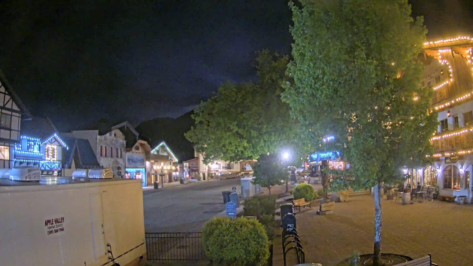A nighttime view of a town square with several buildings decorated with string lights, under a dark, clear sky.