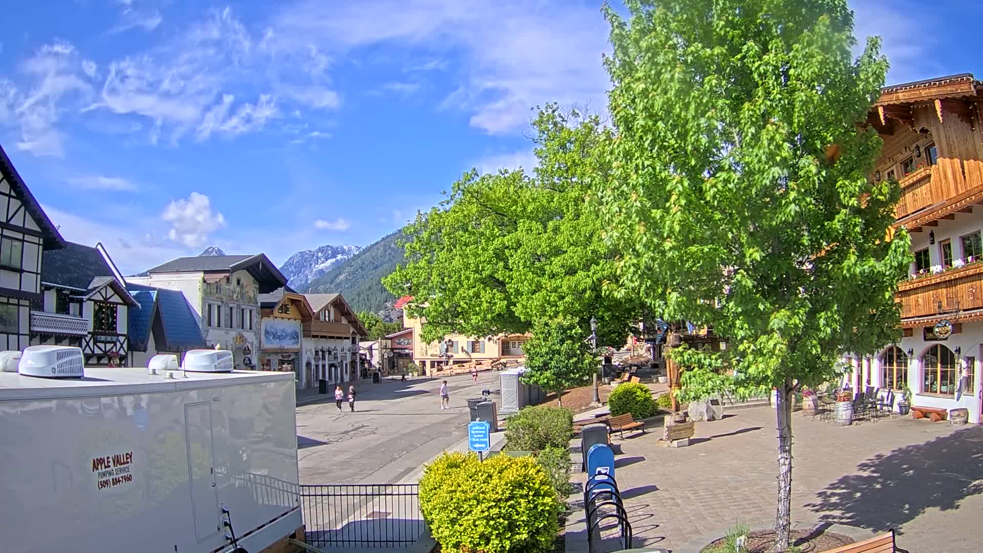 A mostly sunny day reveals a village square with several European-style buildings, lush green trees, and a few pedestrians under a partly cloudy sky, with snow-capped mountains in the background.