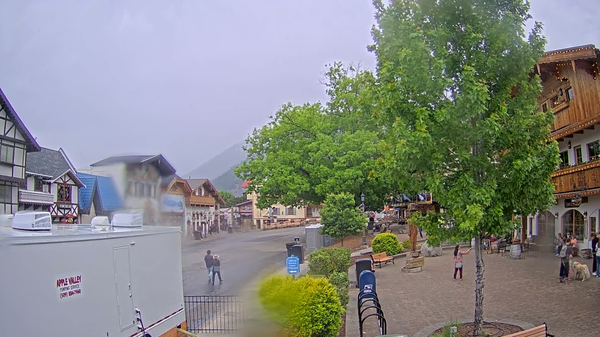A rain-misted town square with European-style buildings, lush green trees, and a few pedestrians is nestled at the base of a mountain.