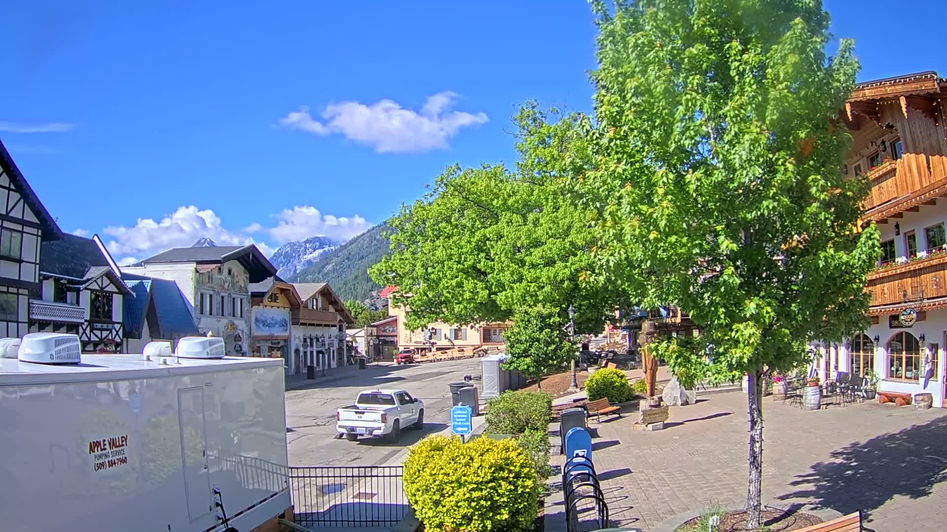 A partly sunny day shows a village square with European-style buildings, lush green trees, and a few parked vehicles, all nestled against a backdrop of snow-capped mountains.