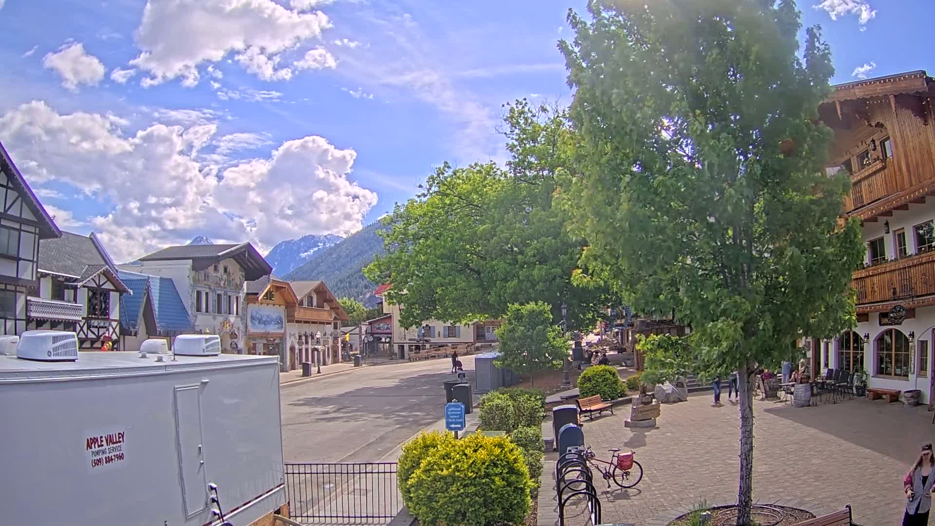 A partly sunny day reveals a quaint village street lined with European-style buildings, lush green trees, and a few pedestrians, all set against a backdrop of mountains.