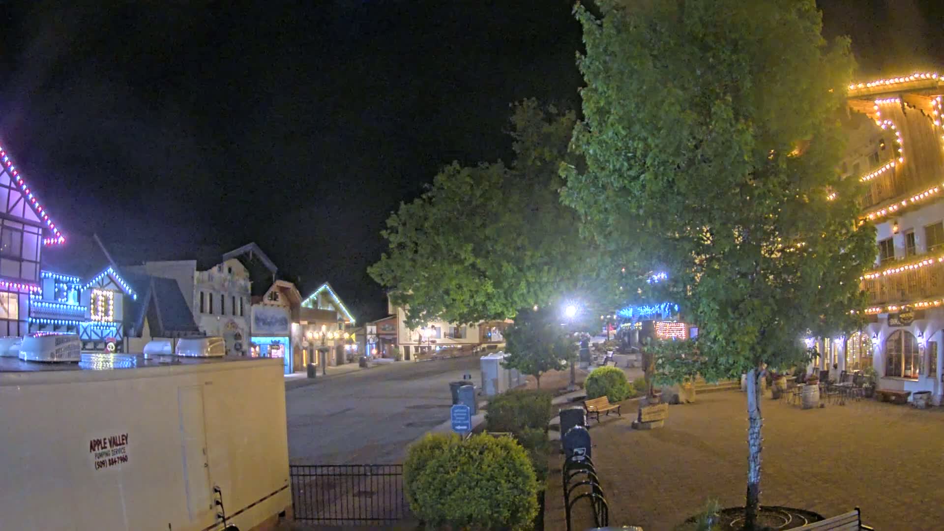 A nighttime view of a town square with several buildings decorated with string lights, surrounded by trees and landscaping.