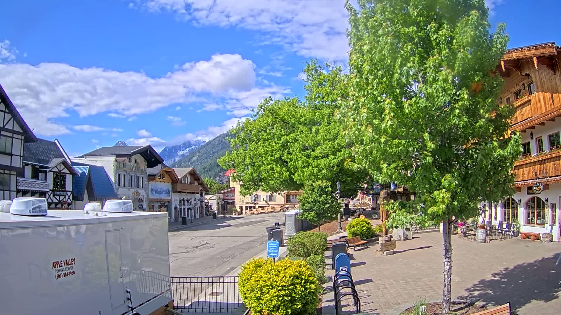 A partly sunny day reveals a quaint town square with European-style buildings, lush green trees, and mountains in the background.