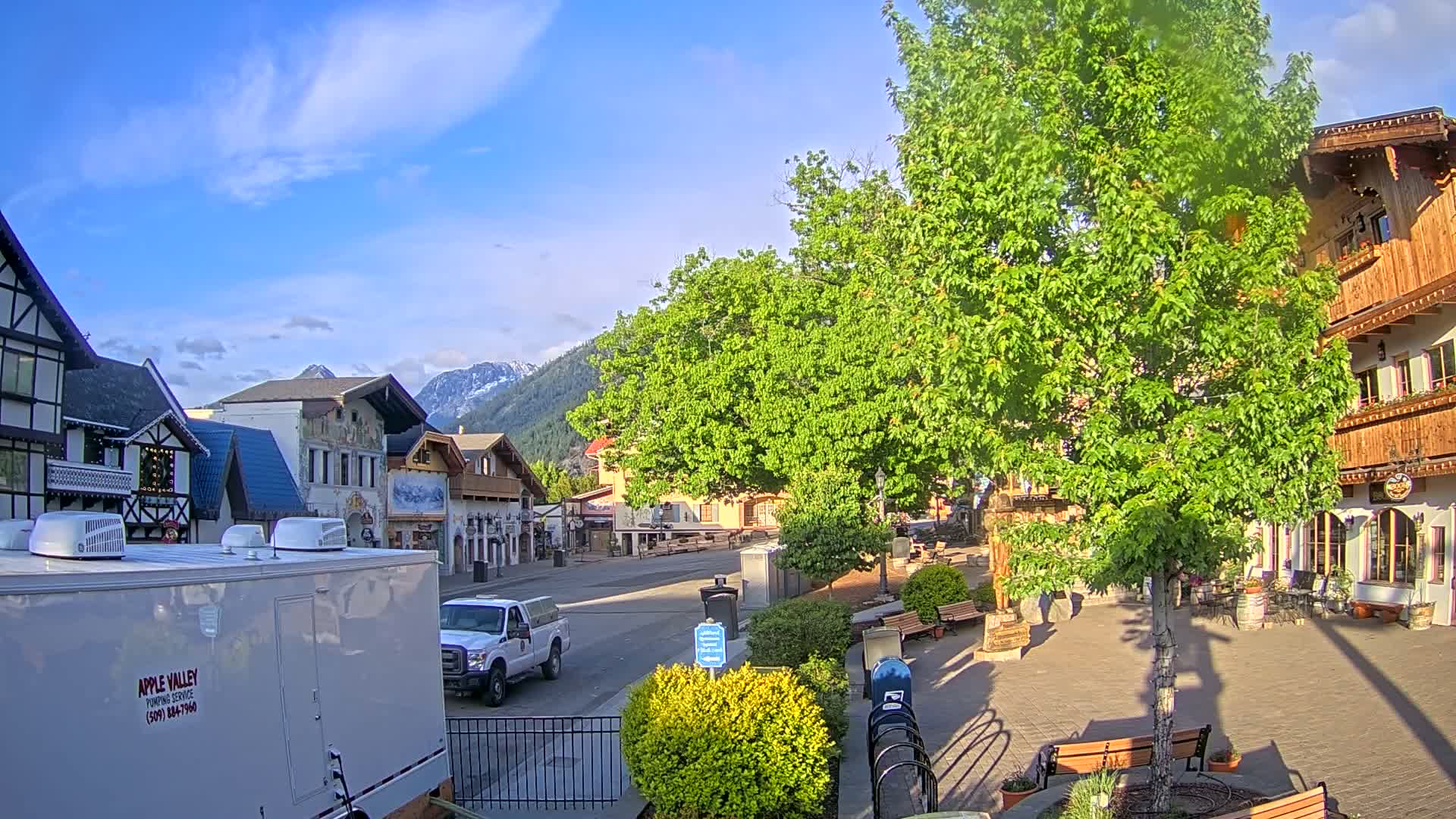 A sunny day reveals a town square with European-style buildings, lush green trees, and a pickup truck parked on a street against a backdrop of snow-capped mountains.
