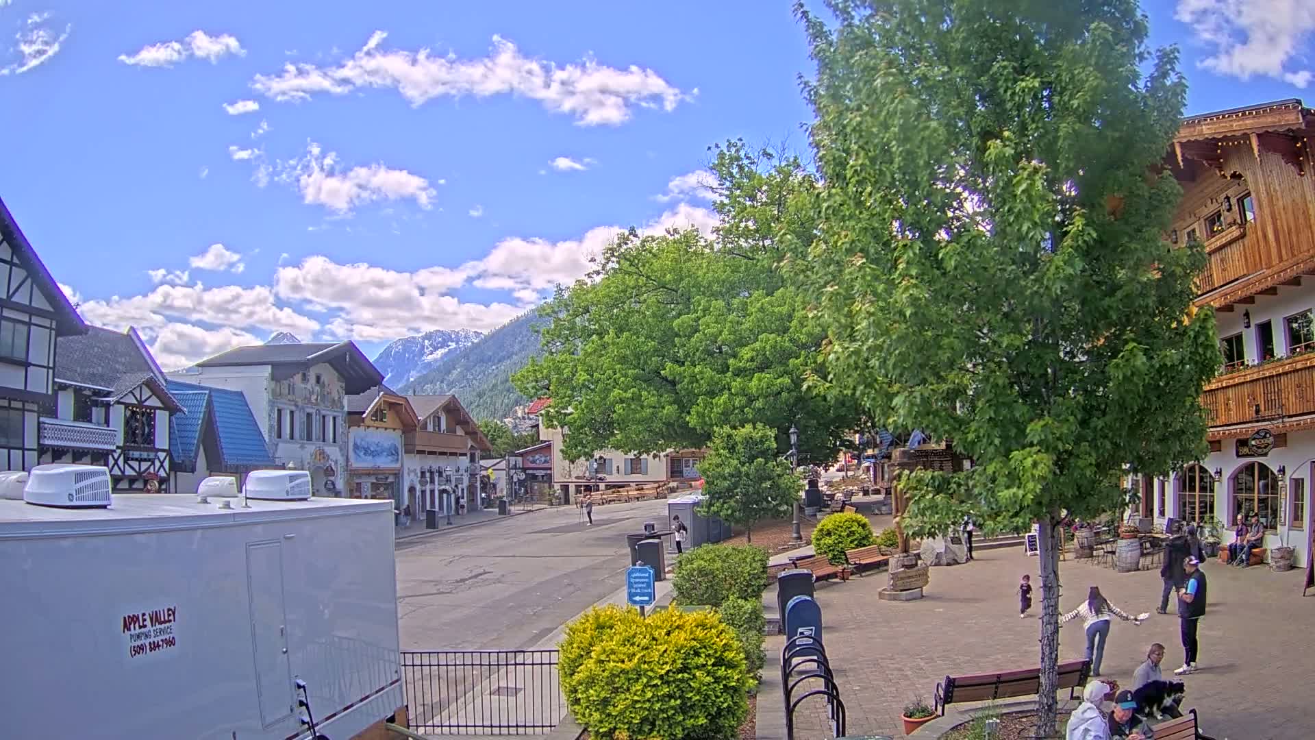 A partly sunny day shows a town square with several people walking around, some sitting on benches, and buildings in a European style, with mountains in the background.