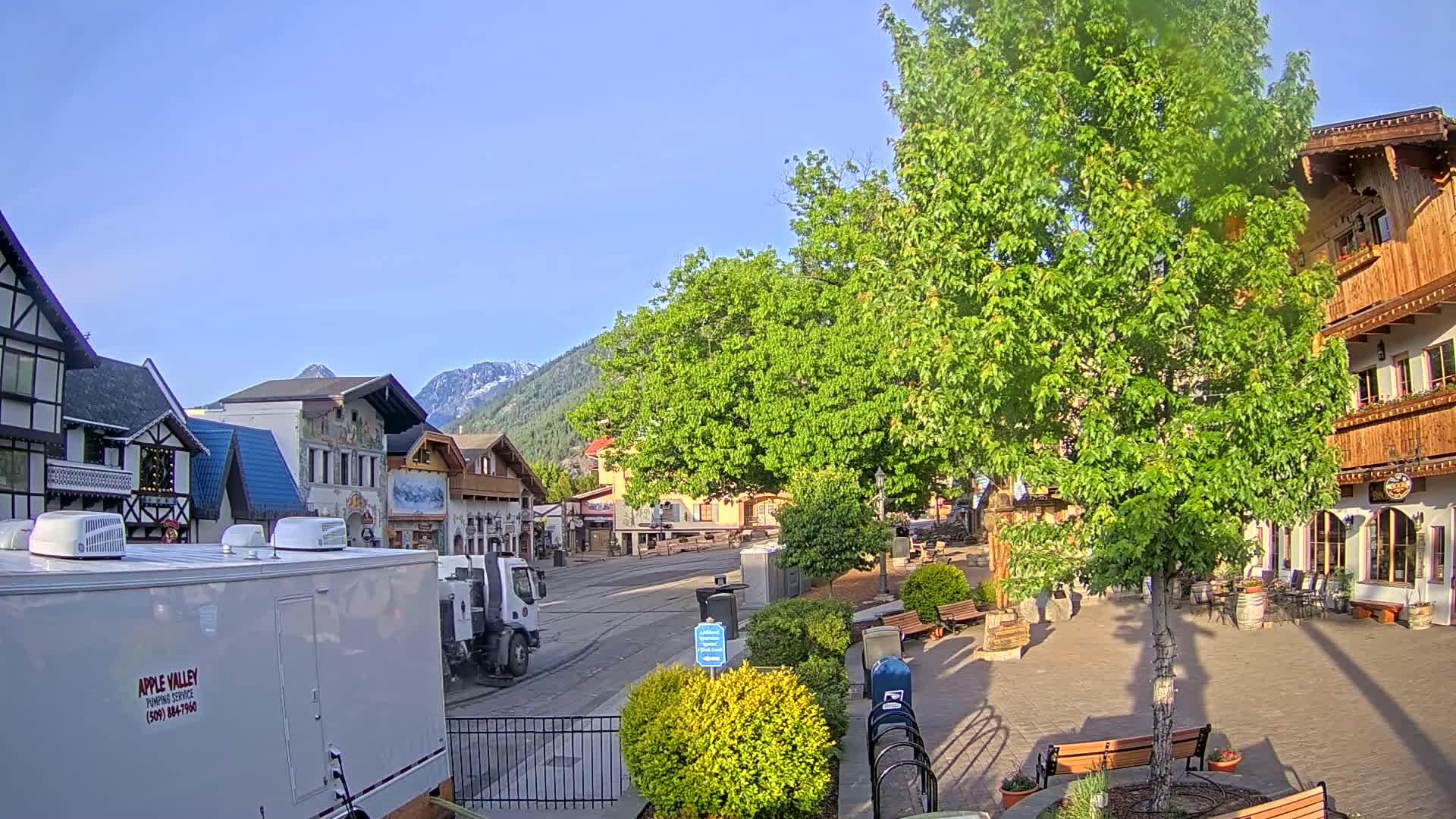 A quaint town square with European-style buildings, lush green trees, and a clear blue sky is seen under sunny conditions.