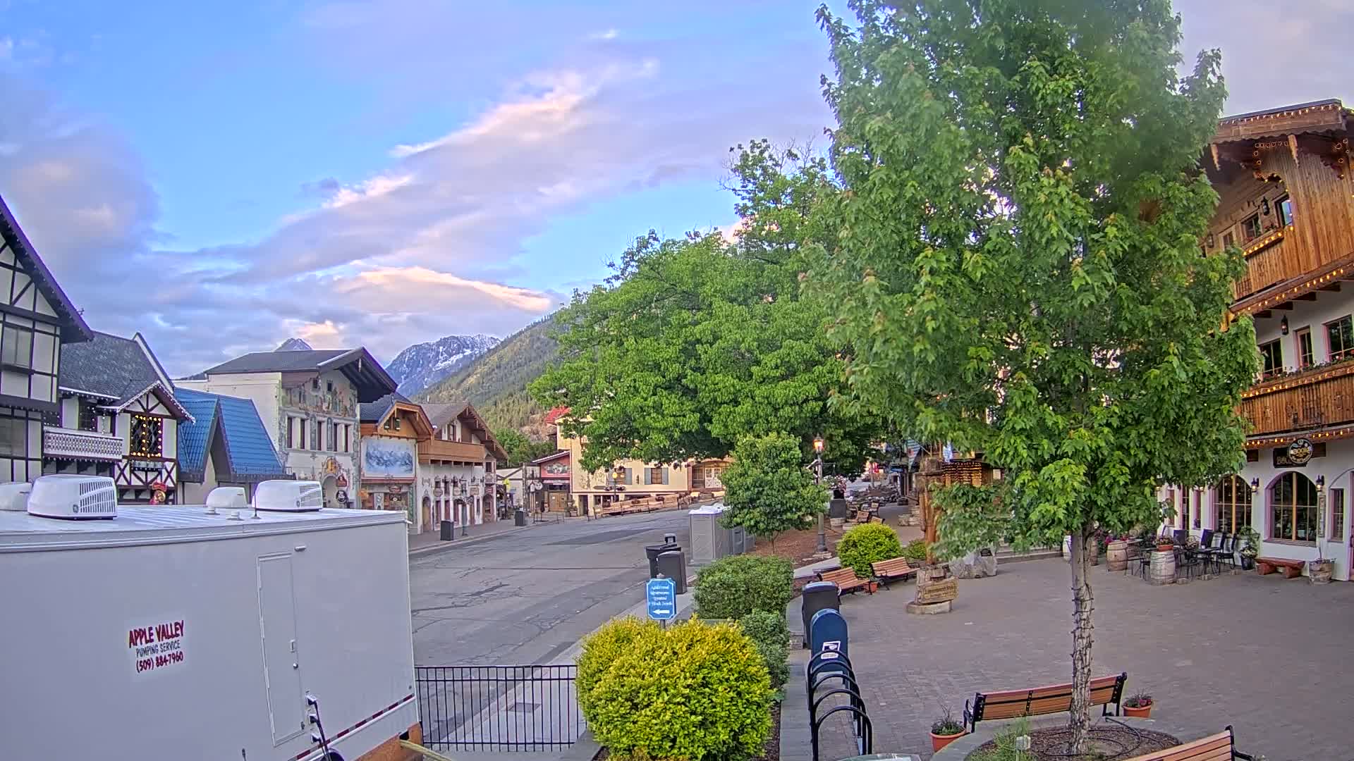 A mostly empty town square with European-style buildings, under a partly cloudy sky and mountains in the background.