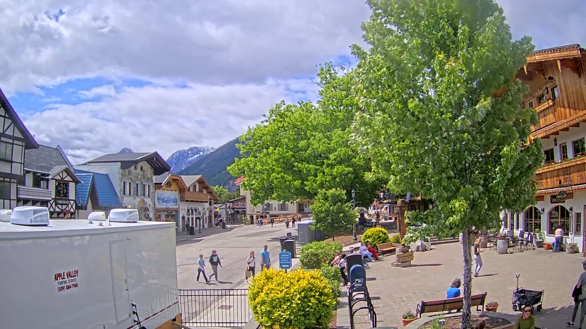 A partly cloudy day reveals a quaint town square with several pedestrians, surrounded by buildings in a Bavarian architectural style, nestled against a backdrop of mountains.