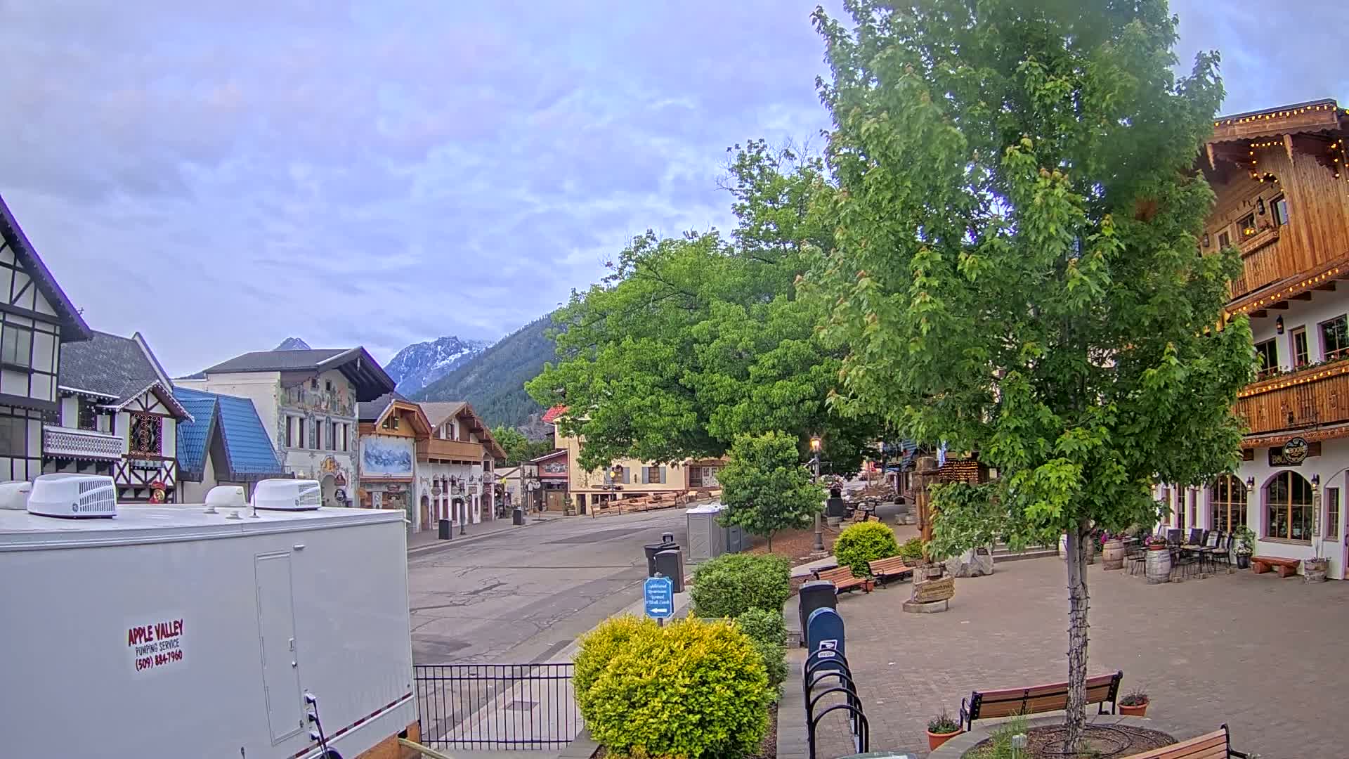 A mostly cloudy day reveals a town square with Bavarian-style buildings, a large tree, and mountains in the distance.