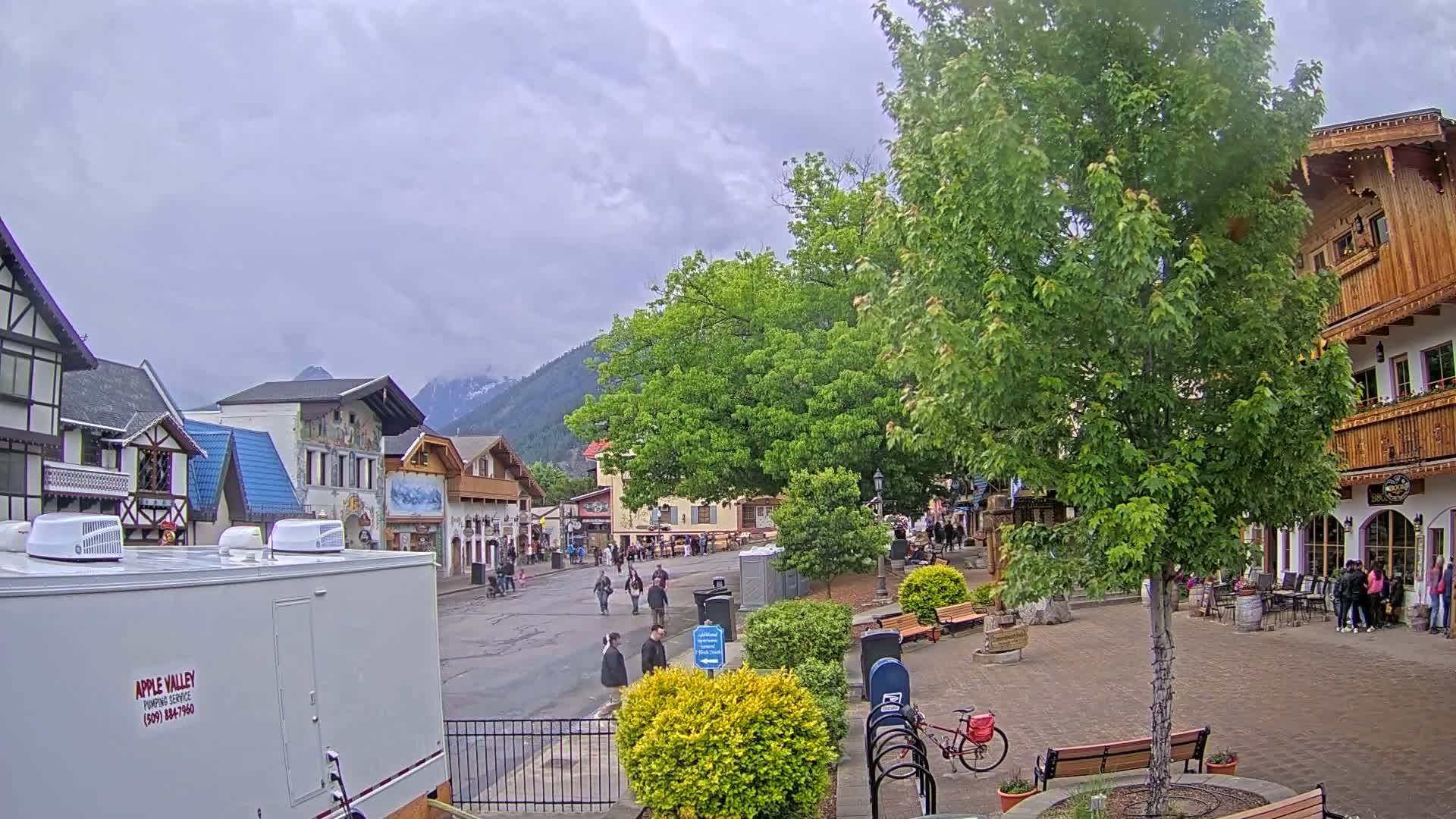 A town square with pedestrians and shops under an overcast sky, nestled against a backdrop of mountains.