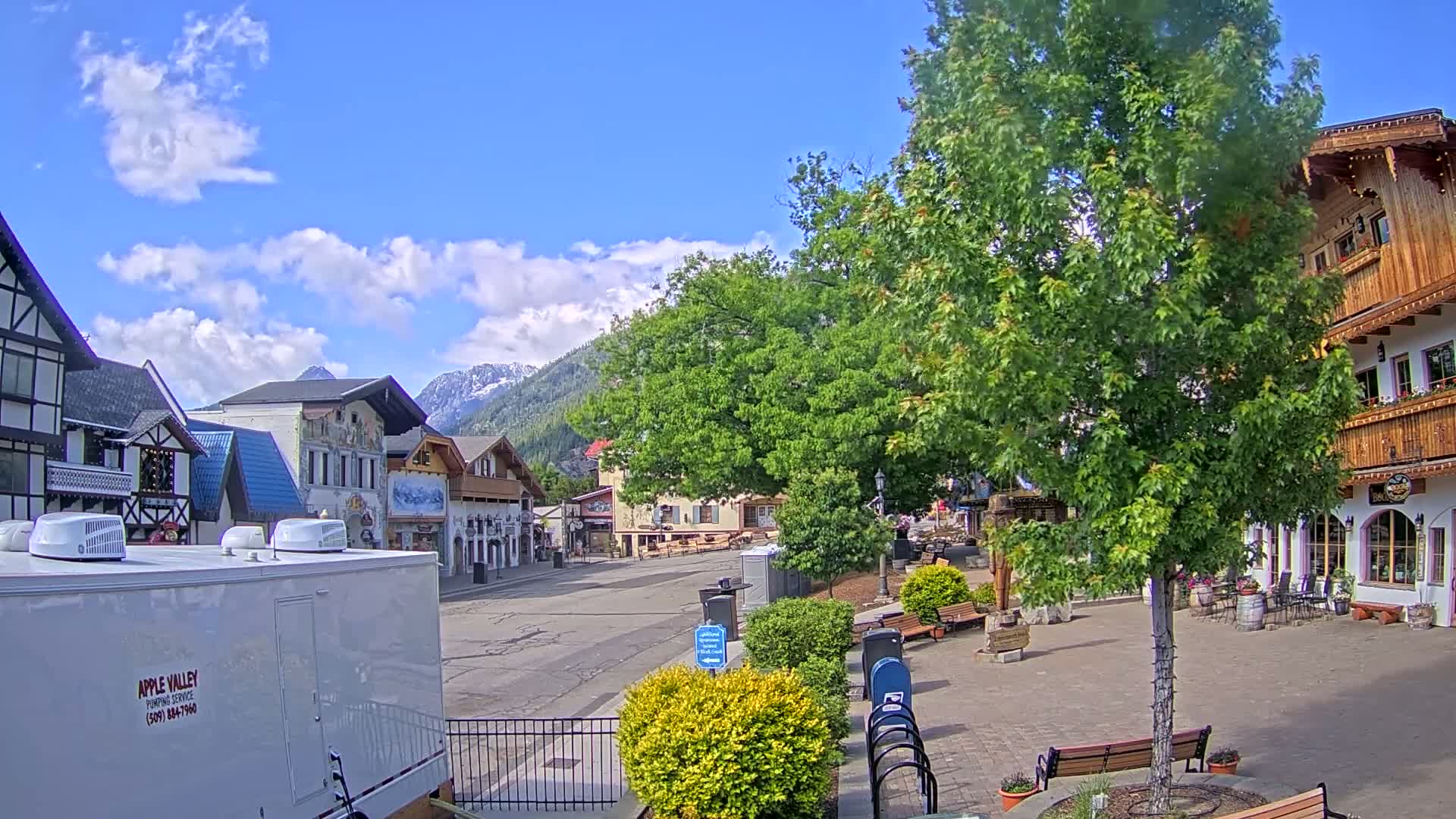 A mostly empty town square with European-style buildings, lush green trees, and mountains in the background under a partly cloudy, sunny sky.