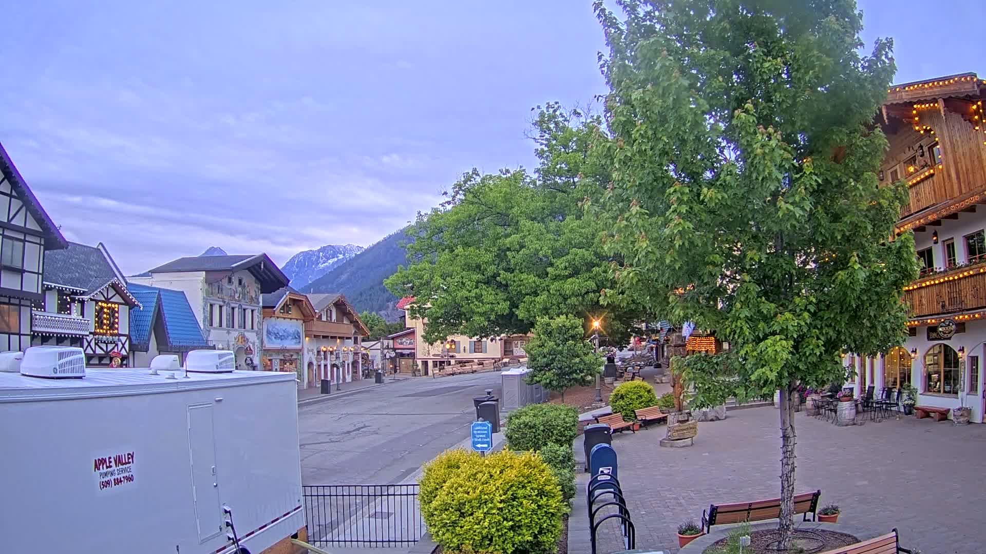 A quaint village street, under a partly cloudy sky, features several European-style buildings nestled against a backdrop of mountains.