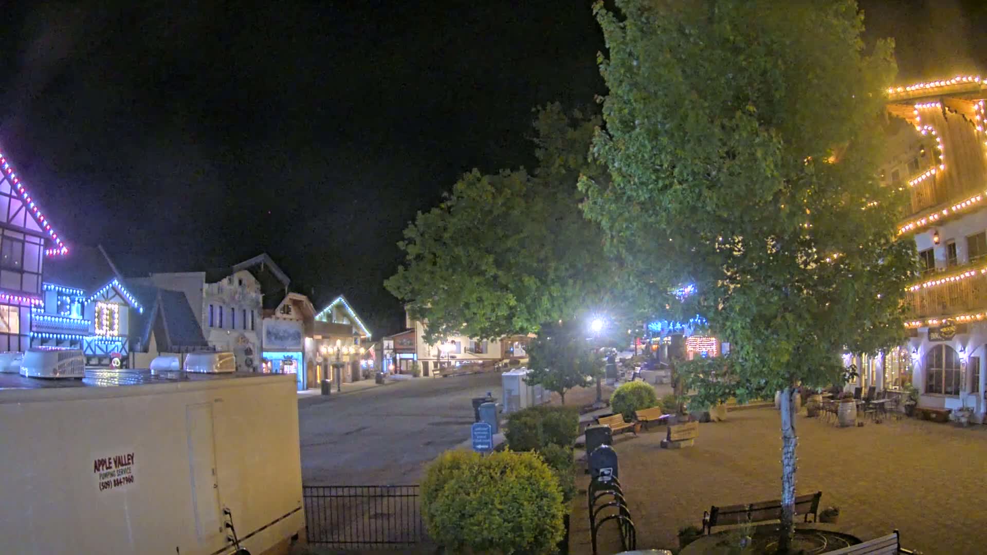 A nighttime scene of a town square with illuminated buildings, trees, and benches under a clear, dark sky.
