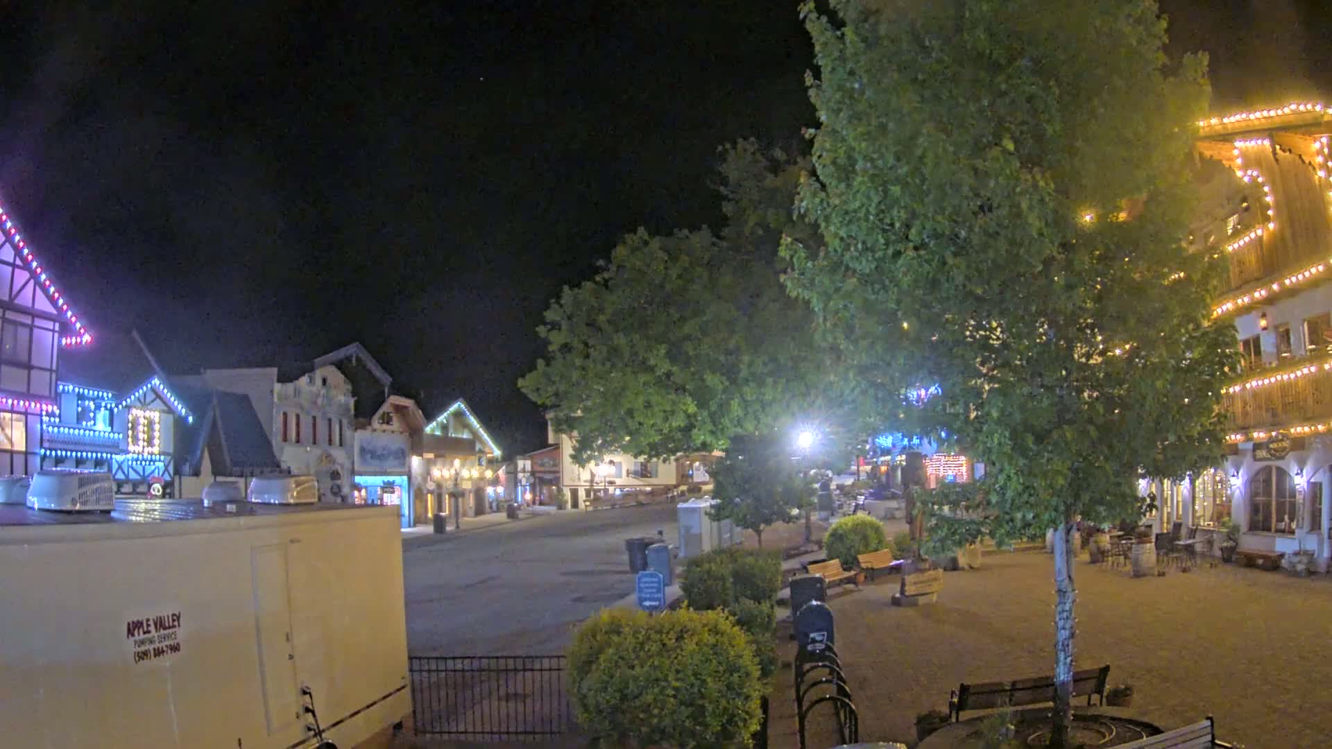 A nighttime view of a town square with several buildings decorated with string lights, trees, and park benches.