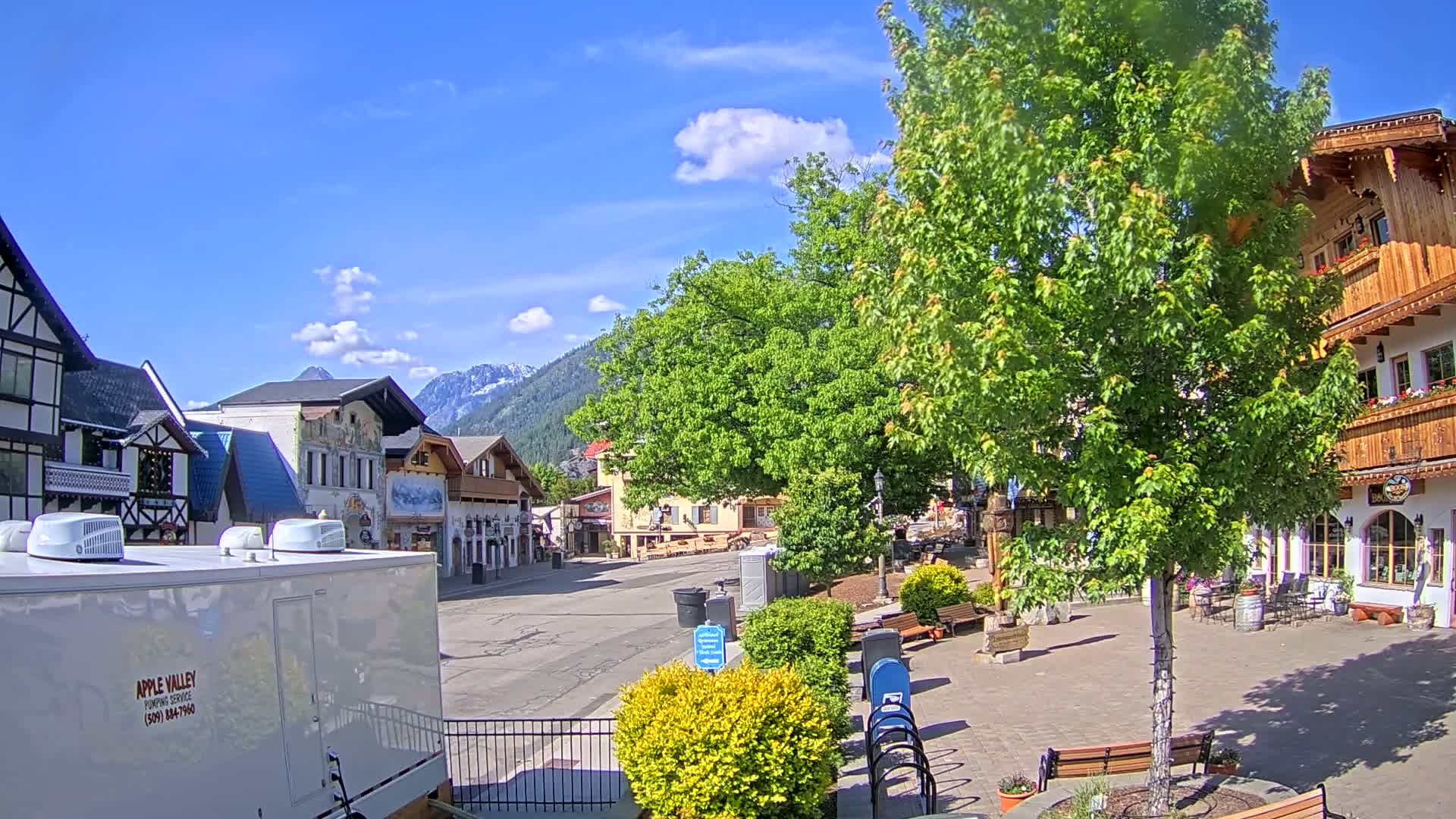 A mostly empty town square with European-style buildings, lush green trees, and mountains in the background under a sunny sky with a few clouds.