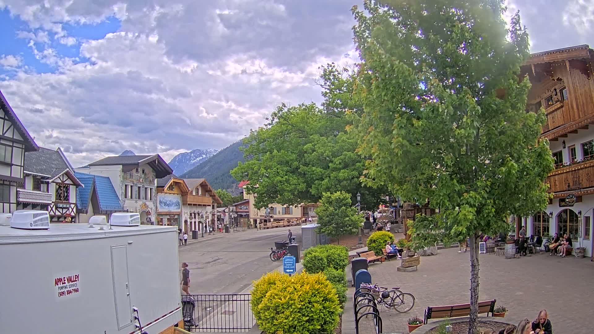A partly cloudy day reveals a quaint village square with European-style buildings, lush greenery, and people enjoying the outdoors, nestled against a backdrop of mountains.