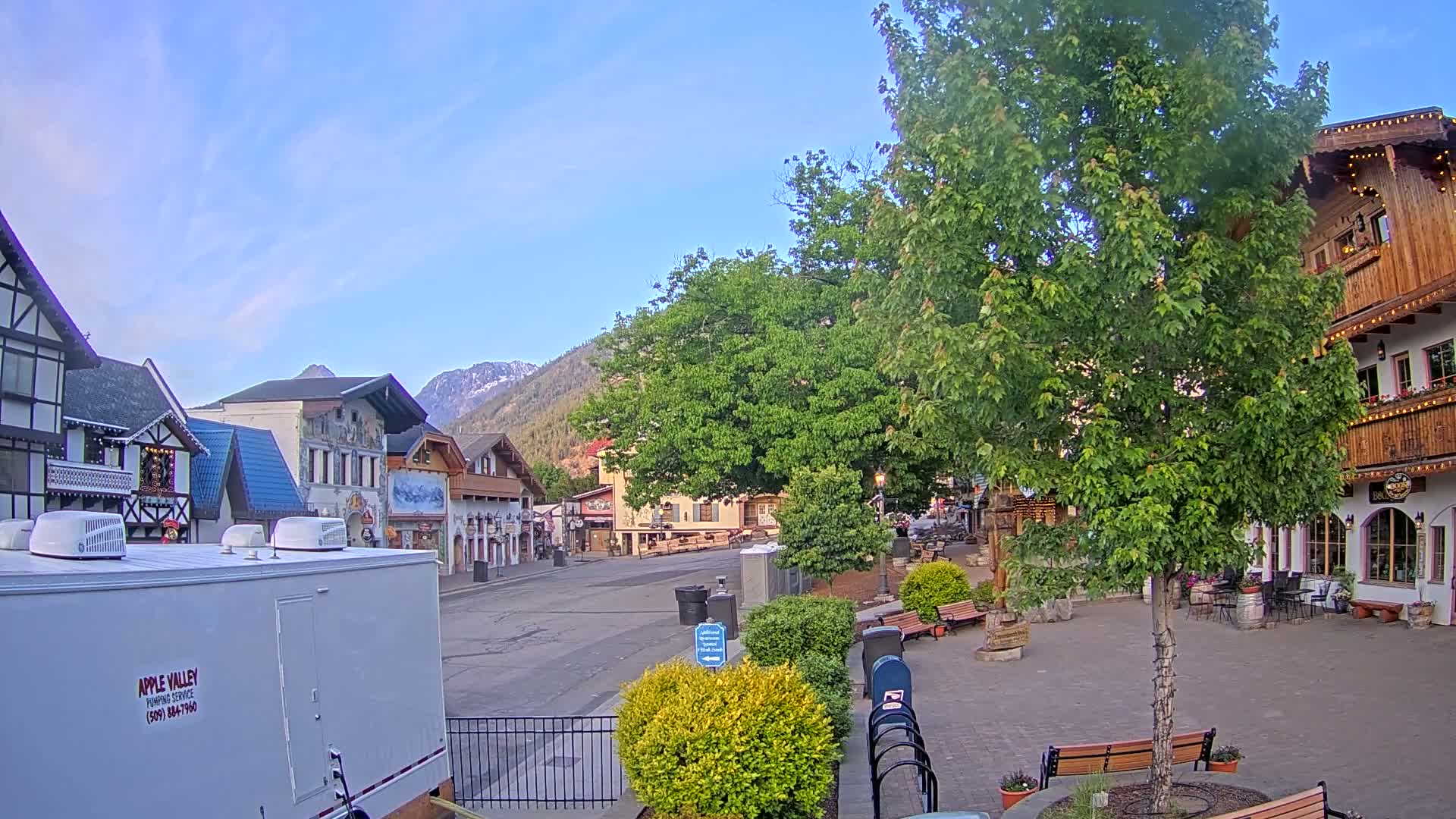 A mostly empty town square with Bavarian-style buildings, under a clear blue sky, is surrounded by lush green trees and mountains in the background.