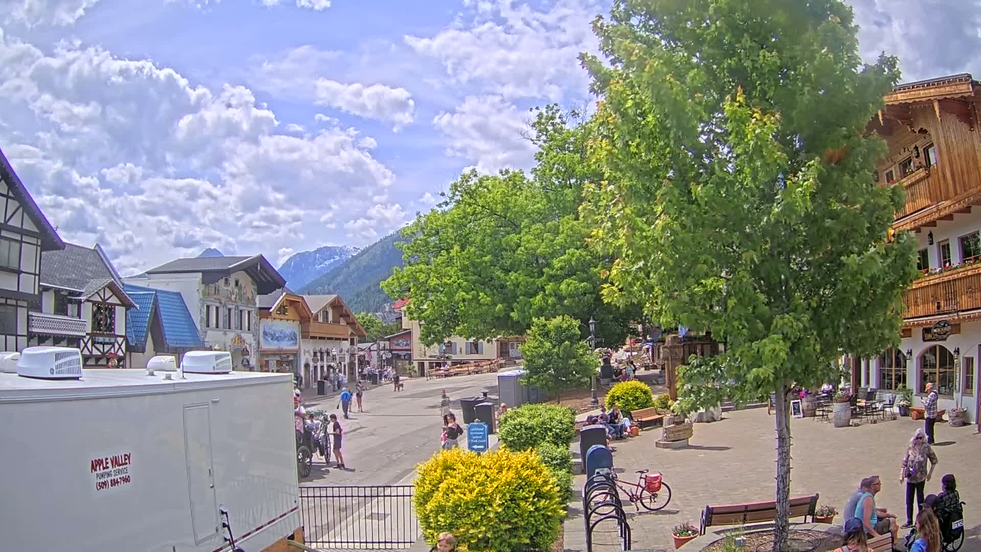 A partly sunny day in a village square with several buildings in an Alpine style, people strolling and sitting, and mountains in the background.