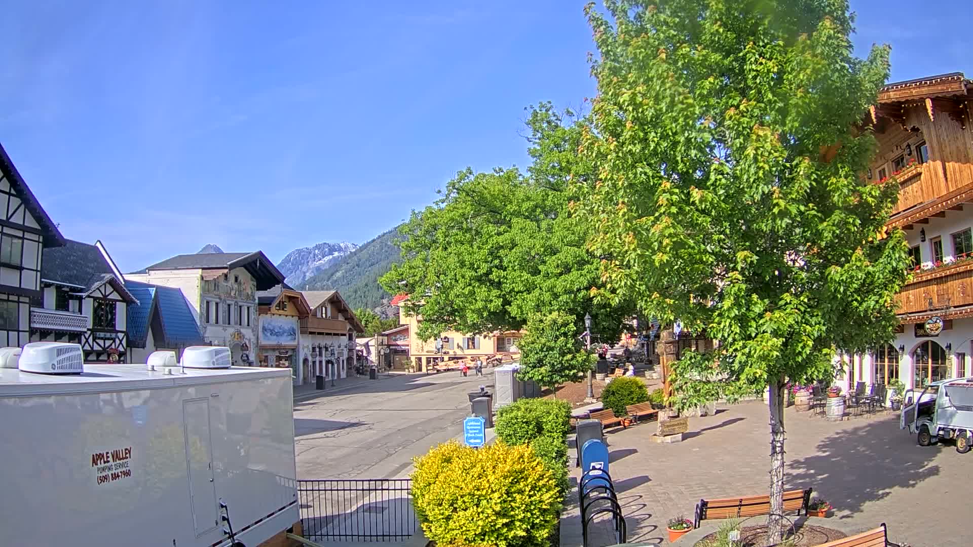 A sunny day in a village square, featuring European-style buildings, lush green trees, and a mountain range in the background.