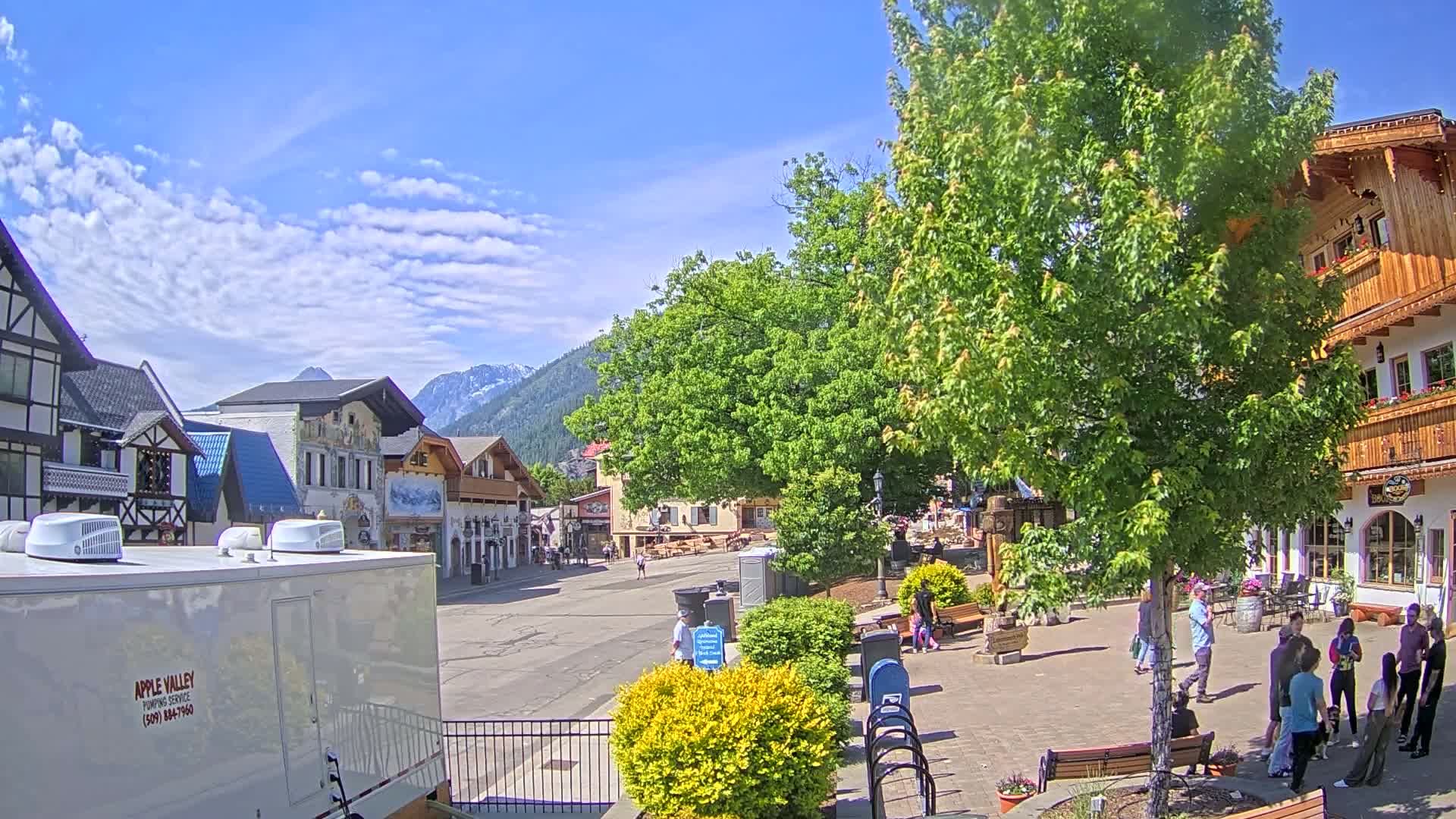 A sunny day reveals a village square with pedestrians, shops, and a large tree, nestled against a backdrop of mountains.
