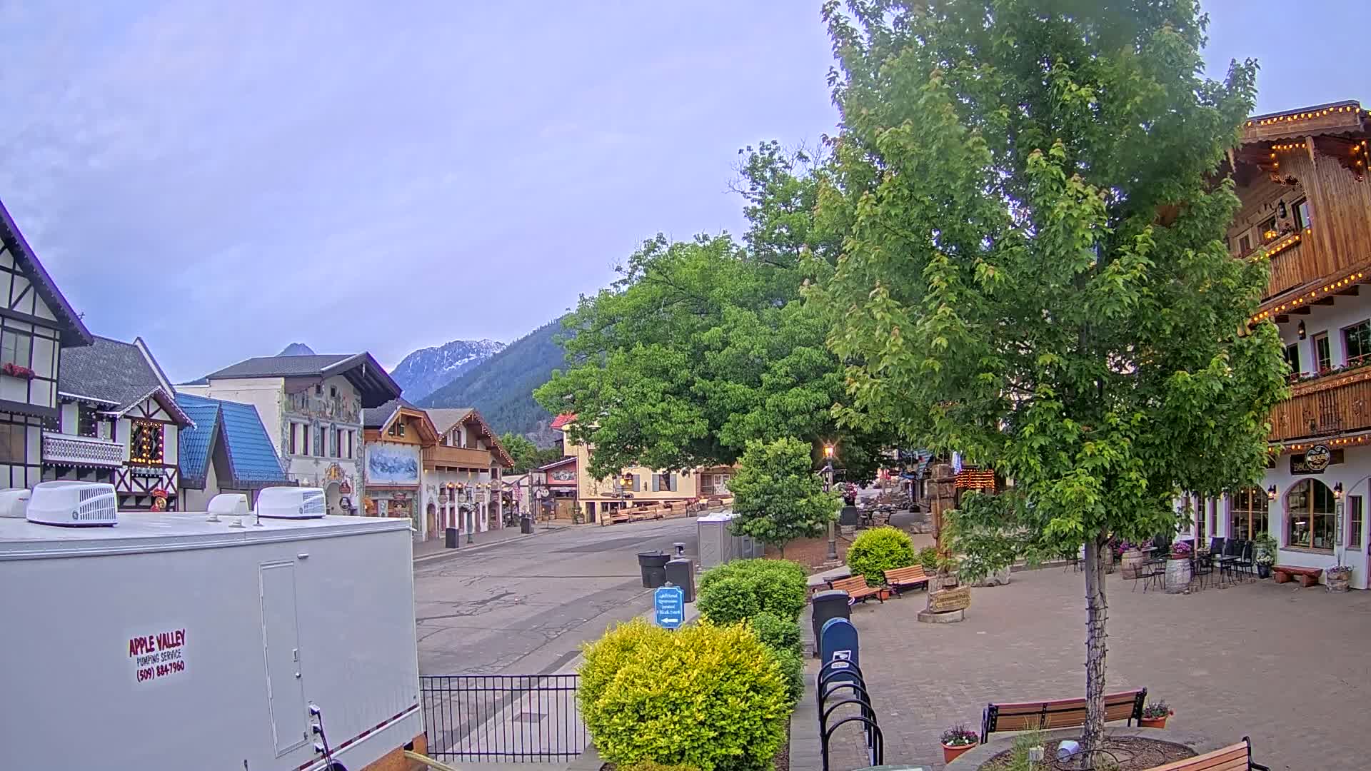 A mostly empty town square with several buildings in a Bavarian style, under an overcast sky, is situated in front of mountains with some snow visible.