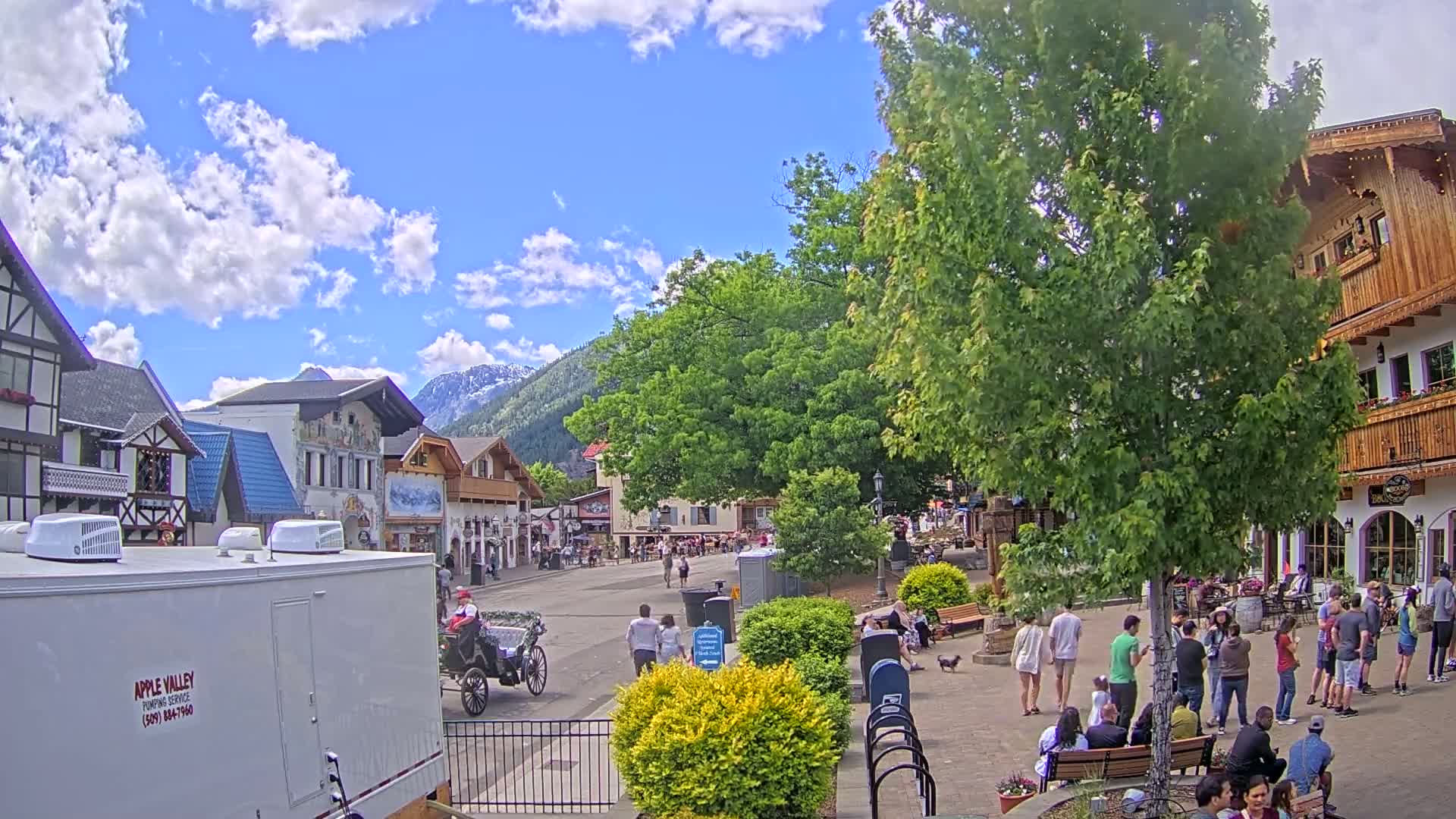 A partly cloudy day in a Bavarian-style village, with people strolling along a street lined with shops and trees, a horse-drawn carriage, and mountains in the background.