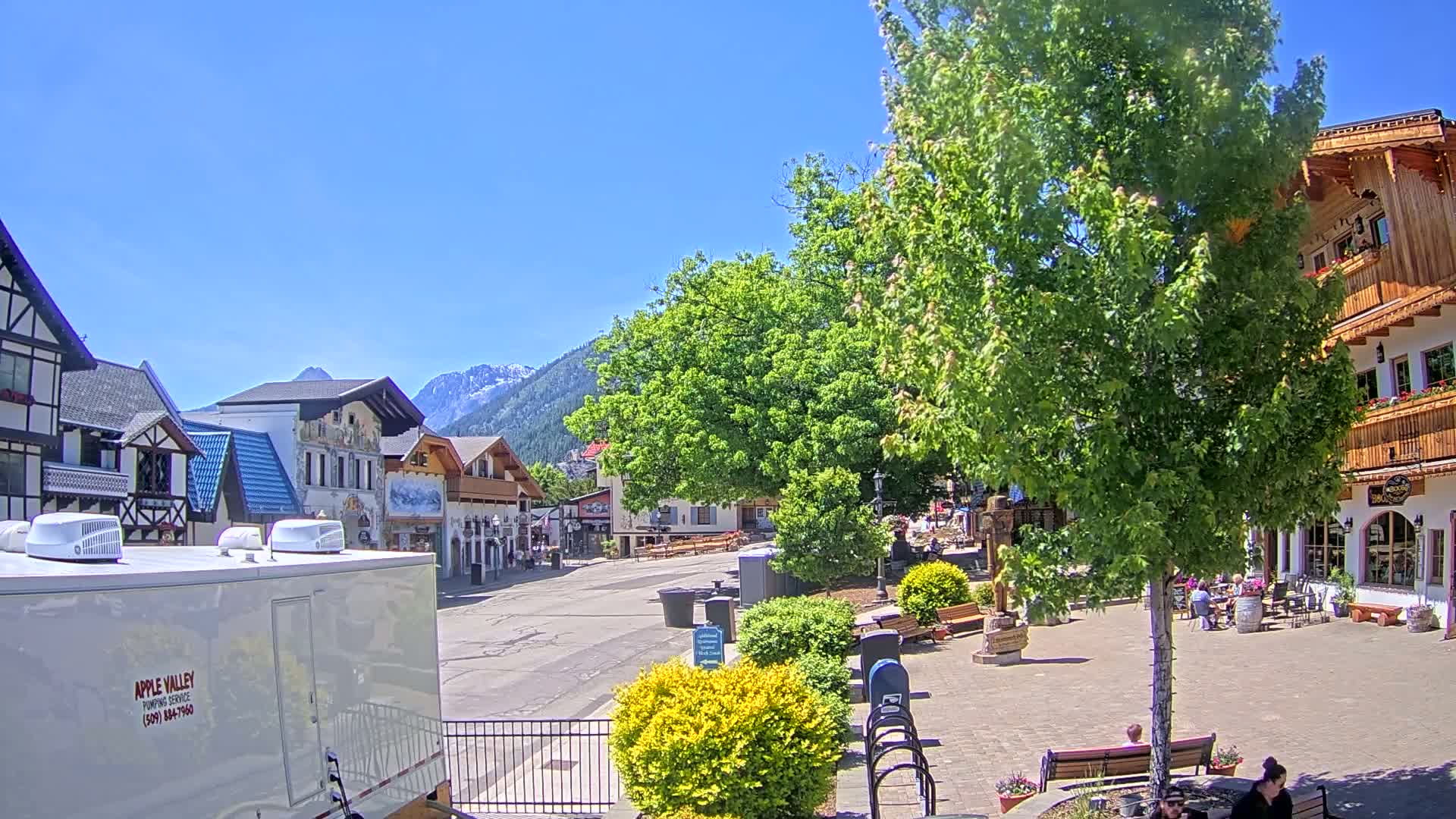 A sunny day reveals a village square with European-style buildings, lush green trees, and several people relaxing on benches, all set against a backdrop of snow-capped mountains.
