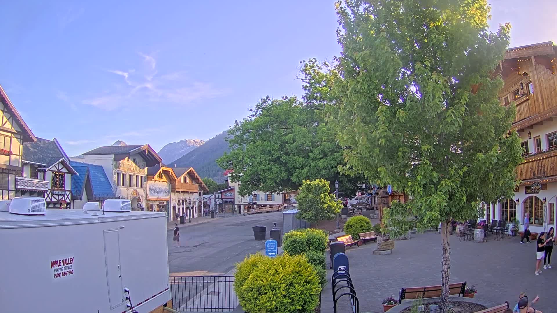 A mostly empty town square with Bavarian-style buildings, under a clear, bright sky, is surrounded by lush green trees and mountains in the background.
